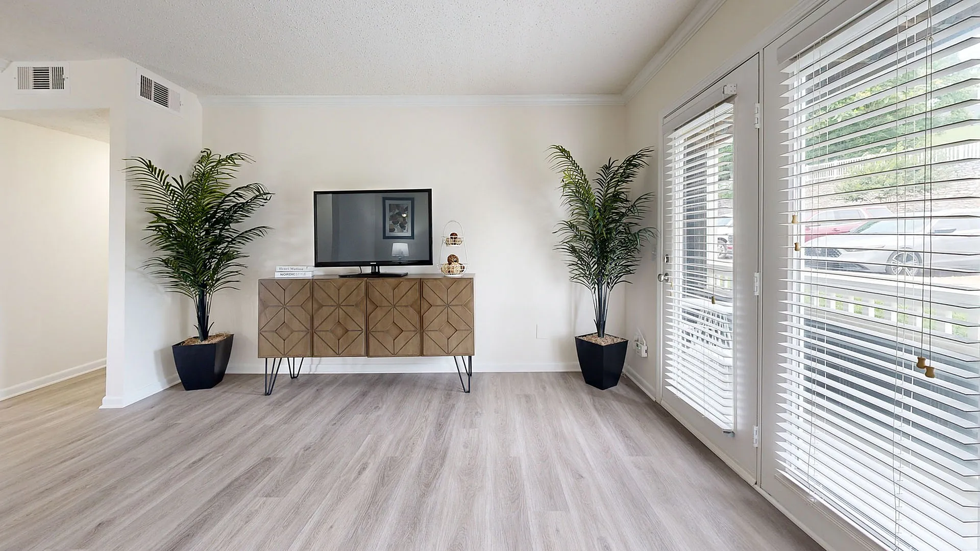 Bright living room with modern decor, featuring a sleek TV stand, elegant potted plants, and large windows with blinds.