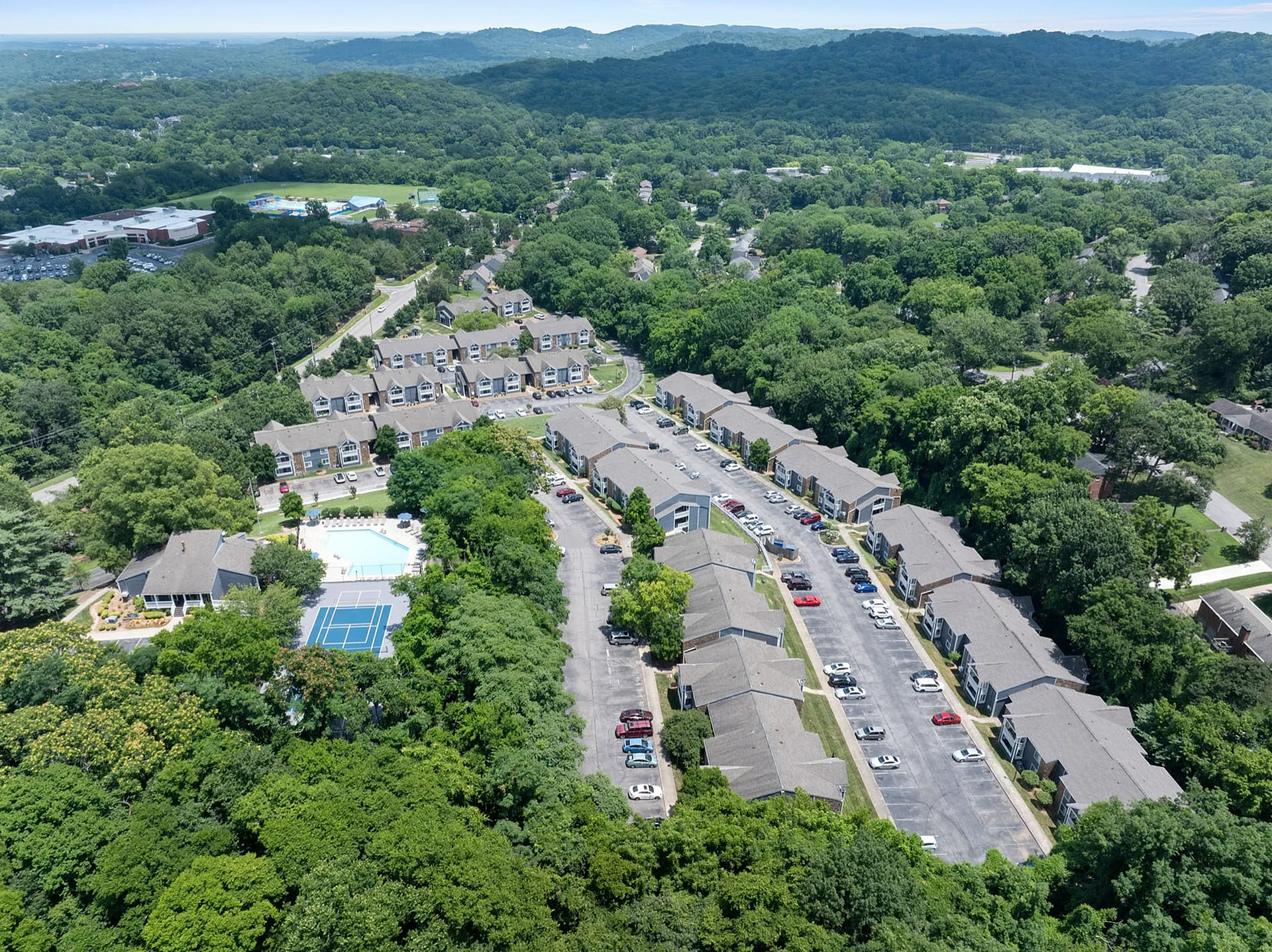 Scenic aerial view of a charming residential community nestled in lush greenery, featuring spacious apartments, a sparkling pool, and a tennis court for a vibrant lifestyle.