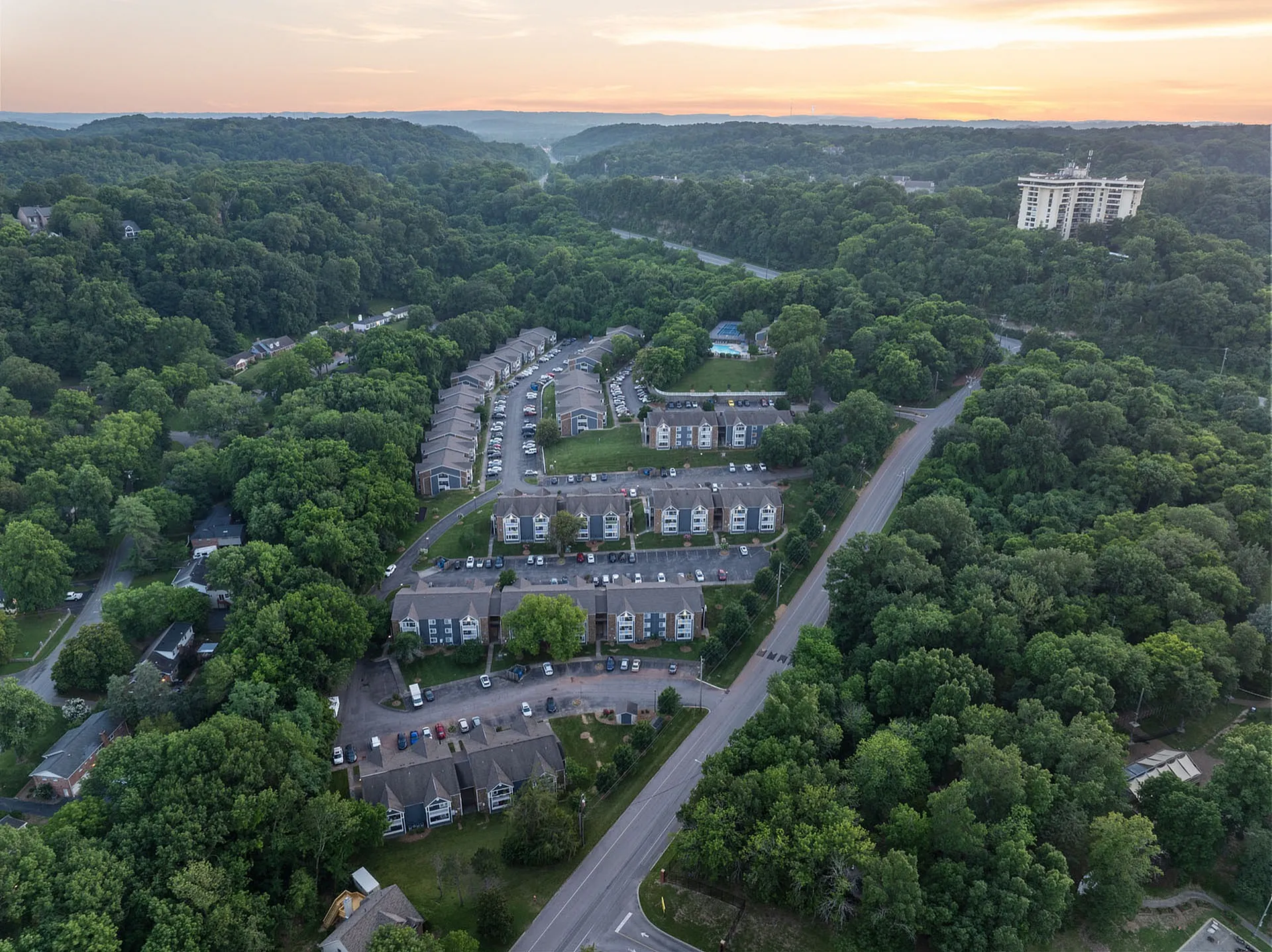 Scenic aerial view of a peaceful apartment complex nestled in lush greenery, offering serene living with ample parking and a swimming pool, framed by a beautiful sunset.