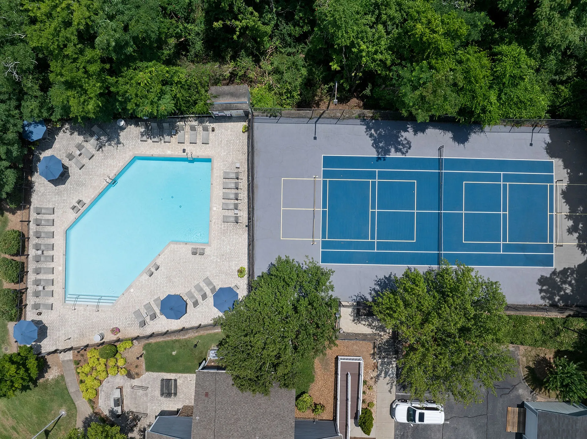 Aerial view of a lush residential complex featuring a sparkling swimming pool surrounded by lounge chairs and umbrellas, adjacent to a well-maintained tennis court, set amidst vibrant greenery.