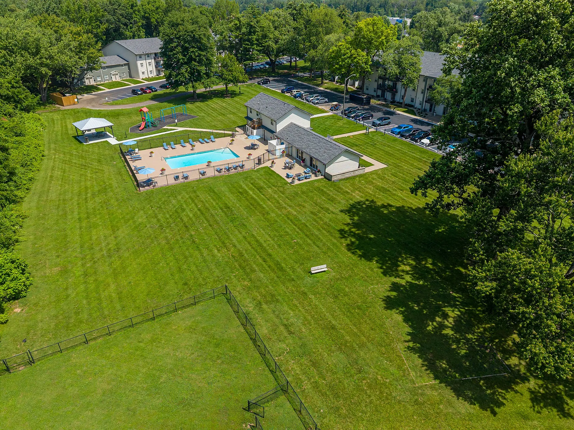 Expansive aerial view showcasing a luxurious apartment complex with a pristine swimming pool, playground, and lush green open space, surrounded by serene greenery.