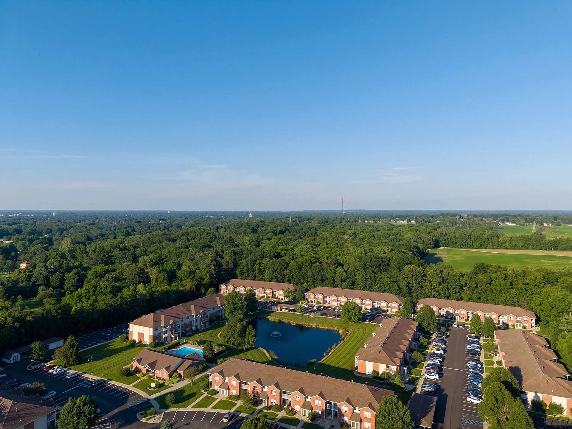 Breathtaking aerial view of a serene apartment complex surrounded by lush greenery, featuring a picturesque pond and a sparkling swimming pool.