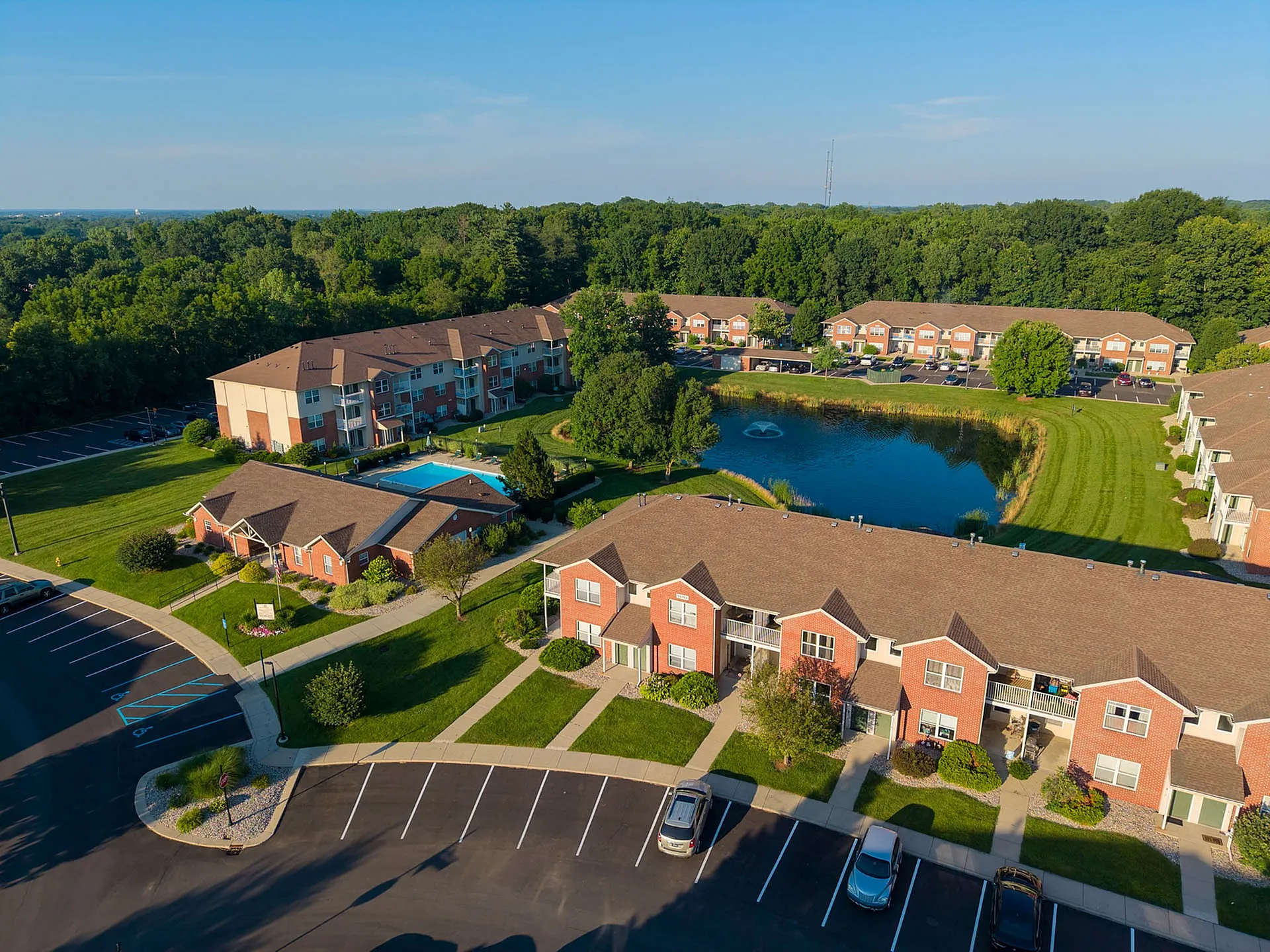 Aerial view of a picturesque apartment community featuring brick buildings, lush green lawns, a sparkling swimming pool, and a serene pond with a fountain, surrounded by wooded areas.
