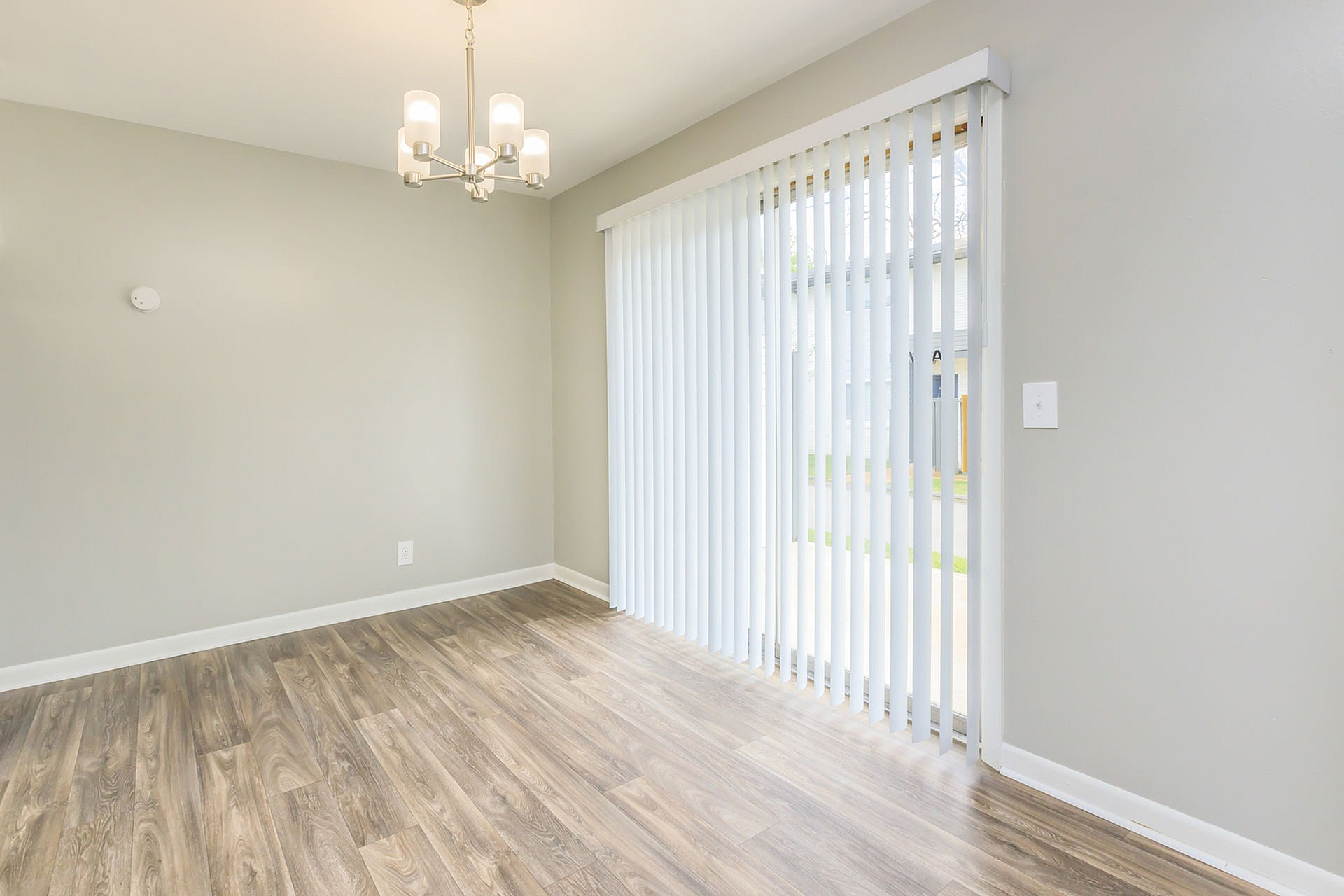 Bright dining area with elegant chandelier and large window draped with vertical blinds, showcasing modern wood flooring.