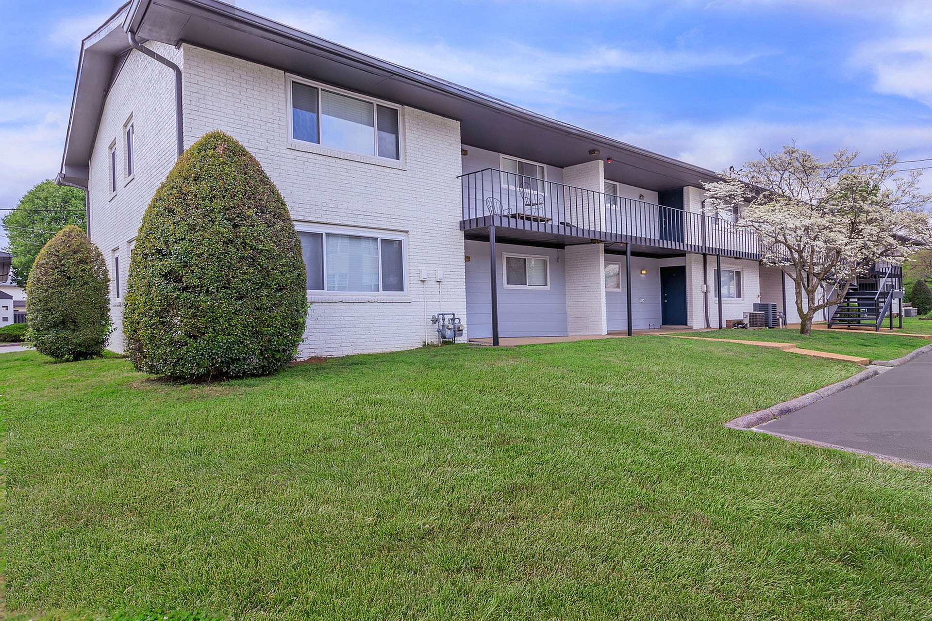 Charming two-story apartment building with white brick facade, lush landscaping, and a welcoming balcony.