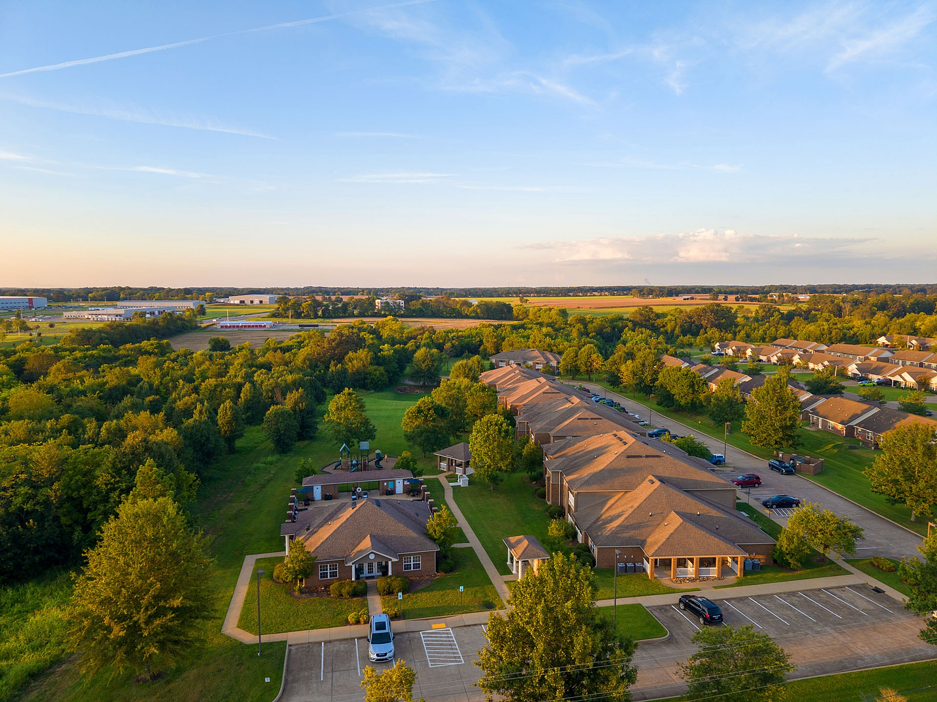 Scenic aerial view of a beautifully landscaped apartment complex surrounded by lush greenery and open fields, offering a tranquil living environment.