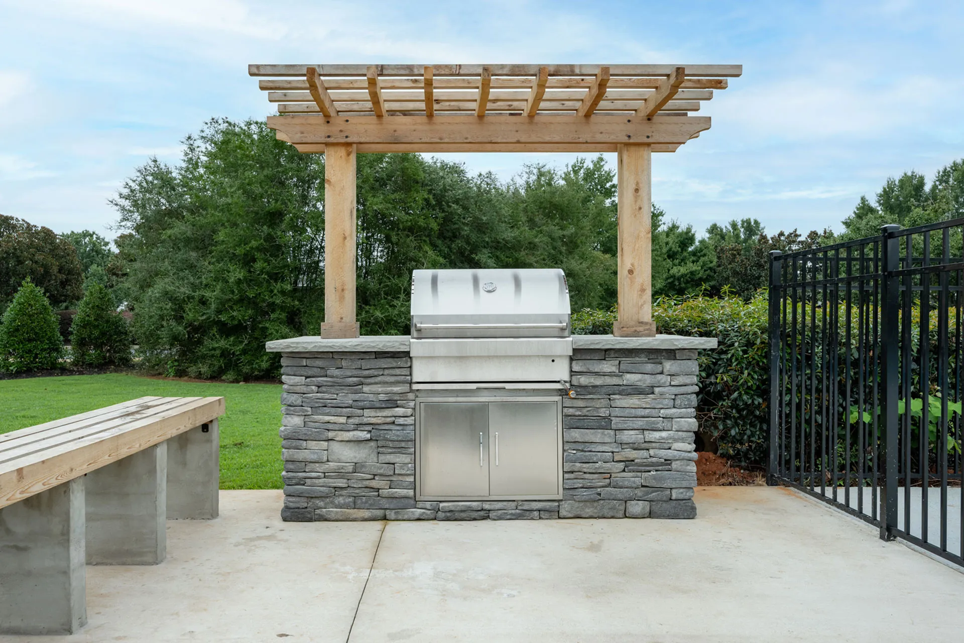 Charming outdoor BBQ area with modern grill and stone seating under a stylish wooden pergola, surrounded by lush greenery.