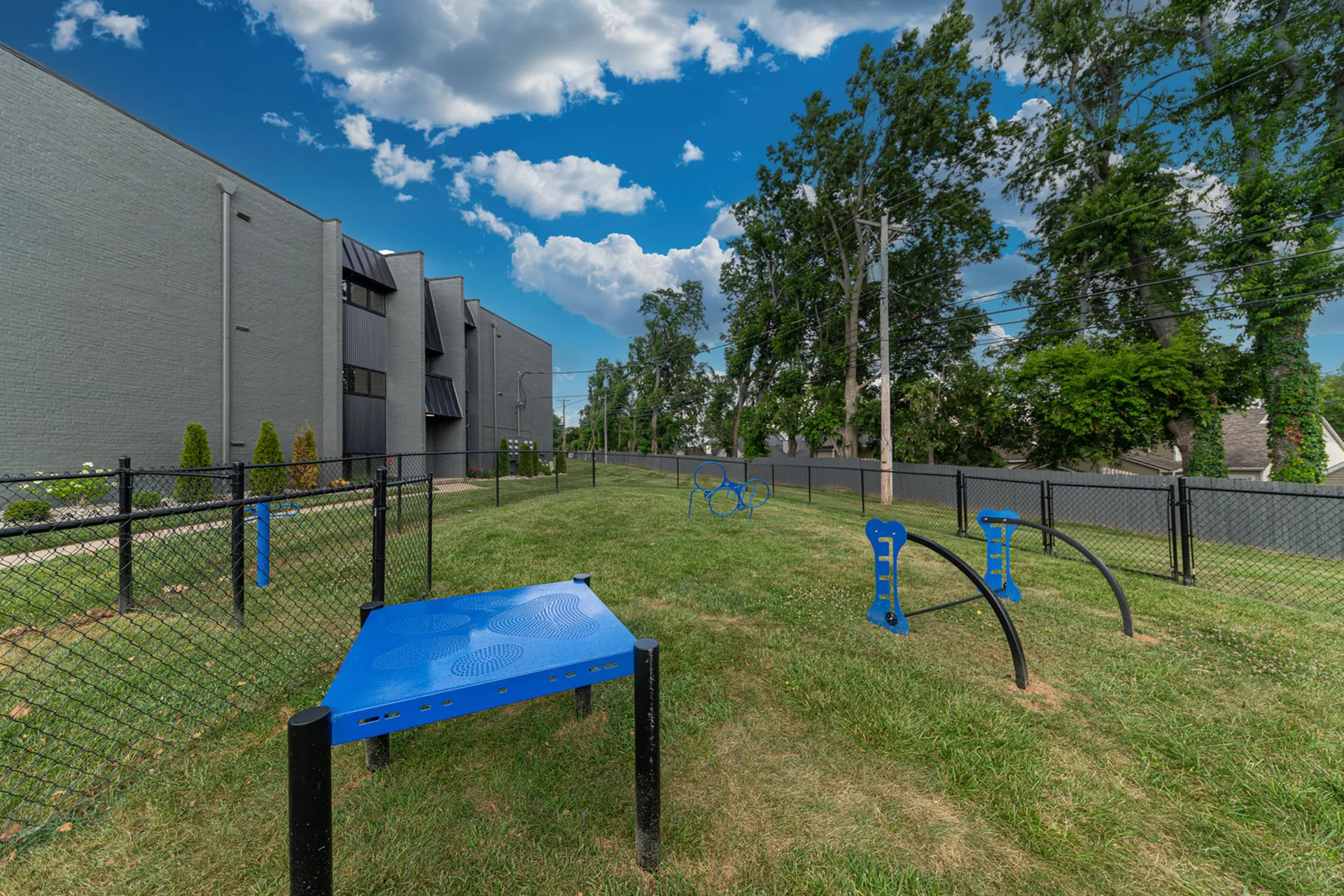 Charming apartment community with a spacious fenced dog park, featuring agility equipment under a clear blue sky.