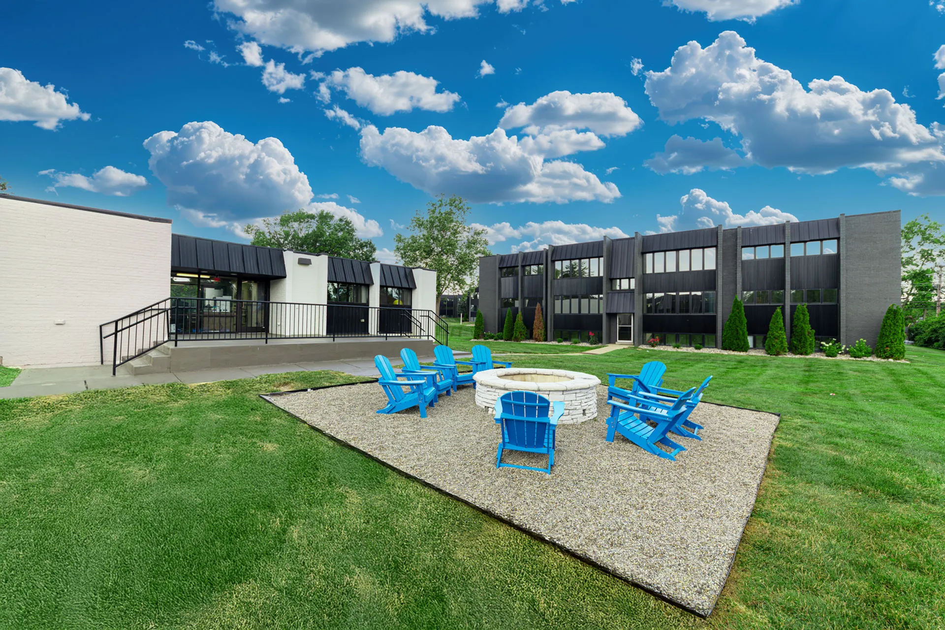 Charming apartment complex with lush green lawns and a cozy outdoor fire pit area featuring blue Adirondack chairs, perfect for relaxation and social gatherings.
