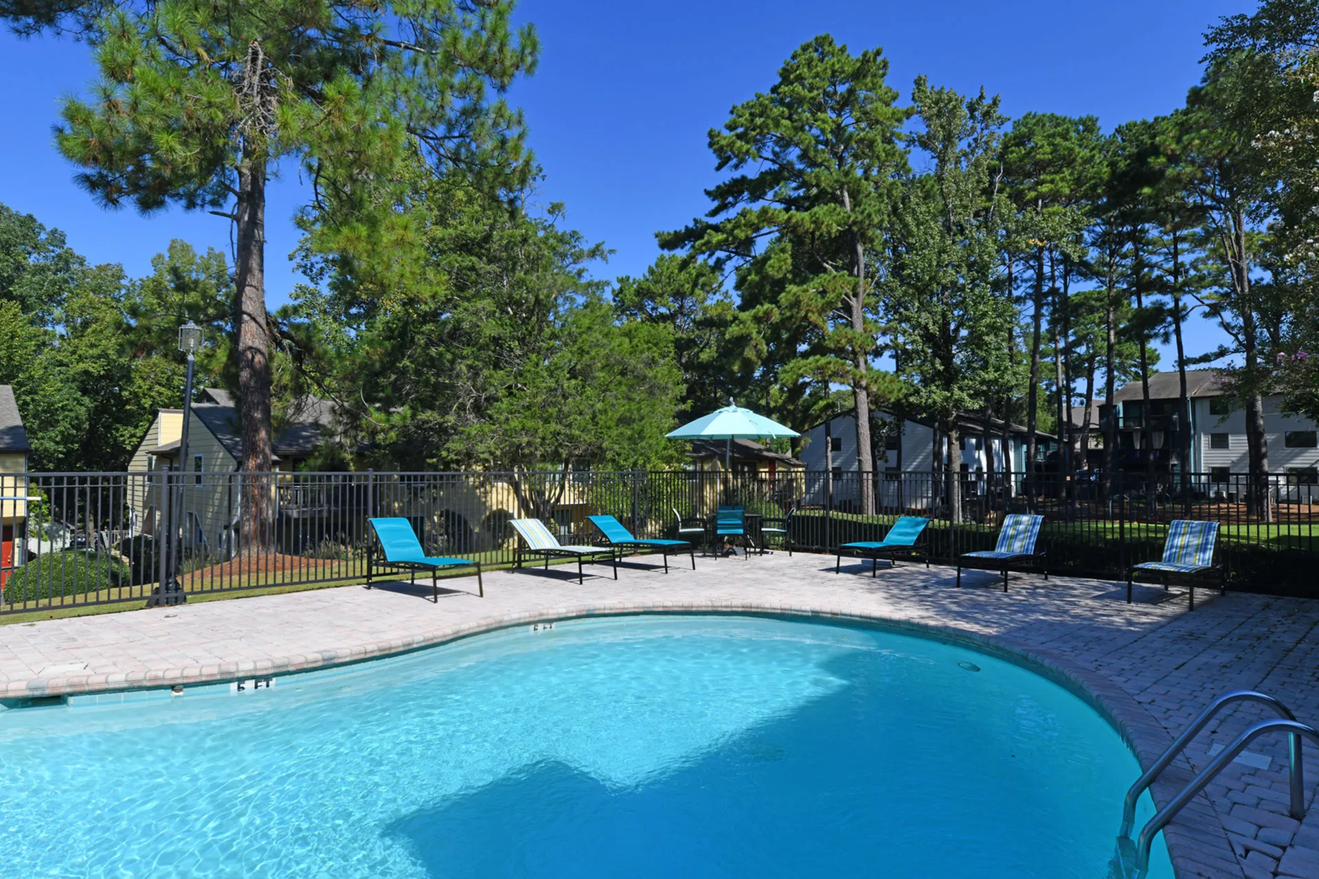 Inviting swimming pool area with vibrant lounge chairs and umbrella, surrounded by lush greenery and modern apartments.