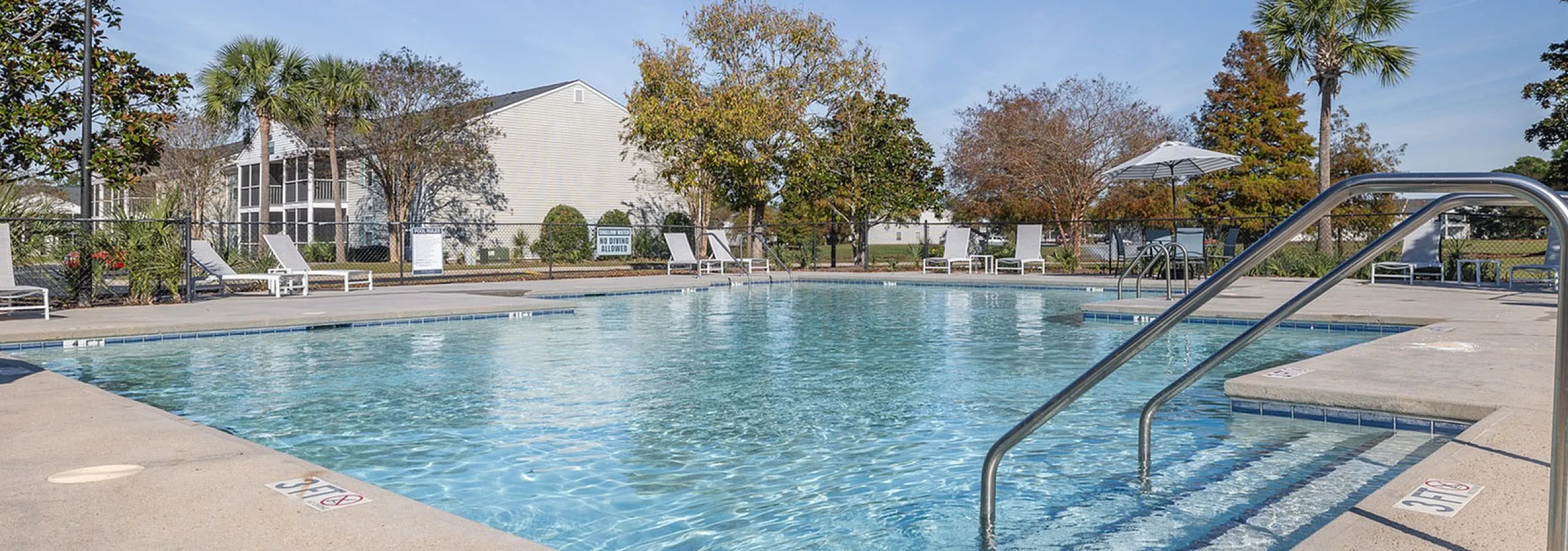 Inviting resort-style pool with poolside lounge chairs, surrounded by lush trees and a clear blue sky.