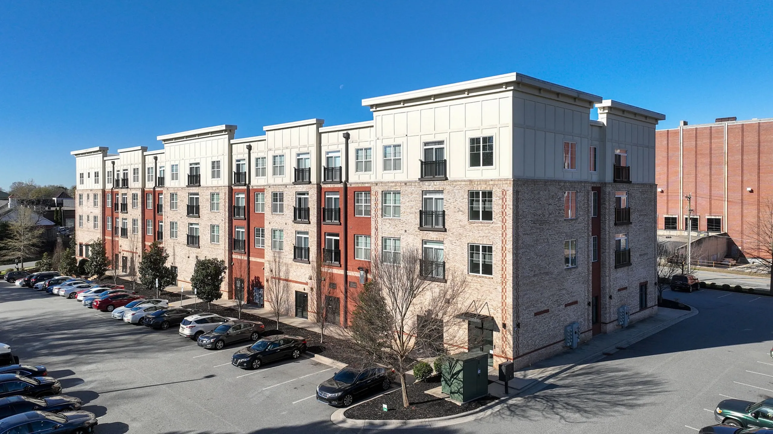 Modern apartment complex with stylish brick and panel facade, ample parking, and vibrant surroundings on a sunny day.