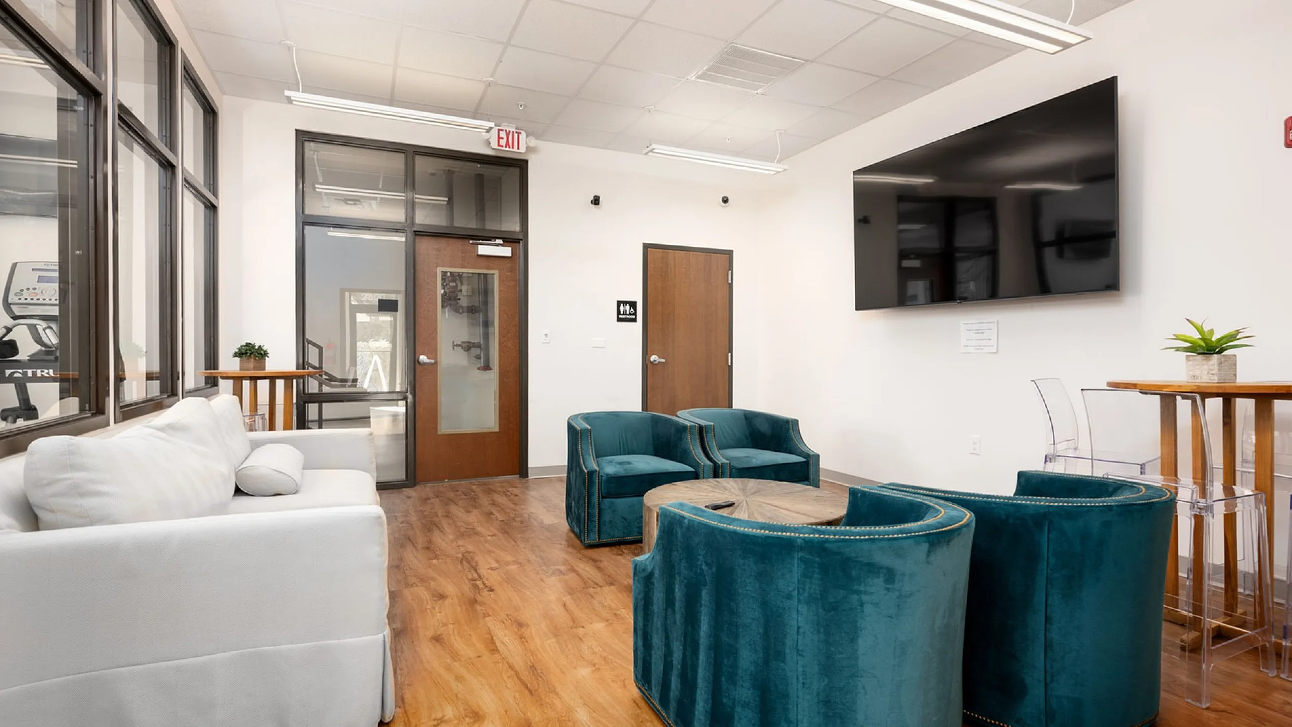 Inviting lounge area with elegant teal chairs, a cozy white sofa, and sleek hardwood floors under a modern wall-mounted TV, perfect for relaxation and social gatherings.
