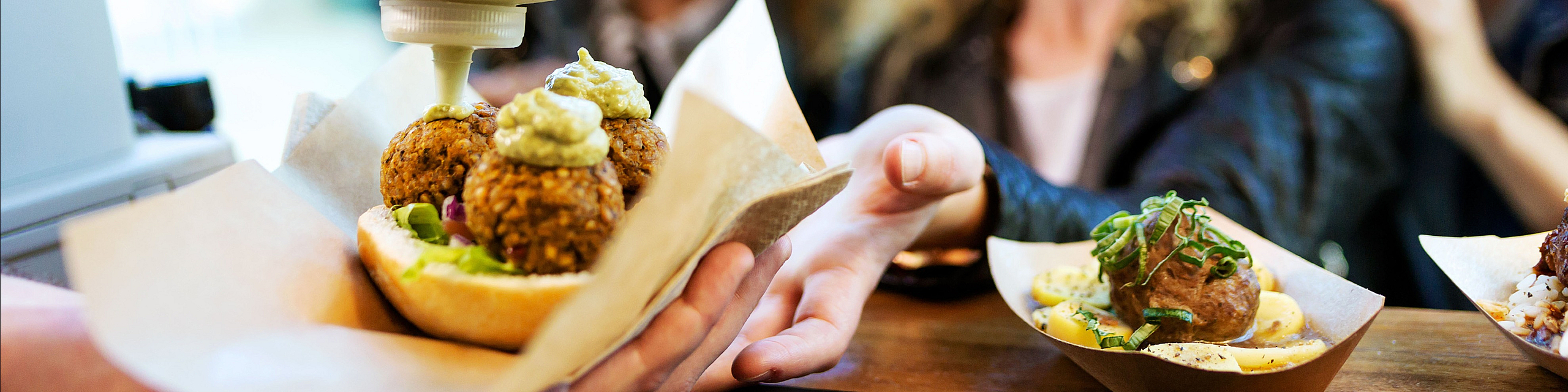 Vibrant scene of friends eagerly receiving delicious gourmet street food, with falafel balls and sauce artfully served in eco-friendly containers.