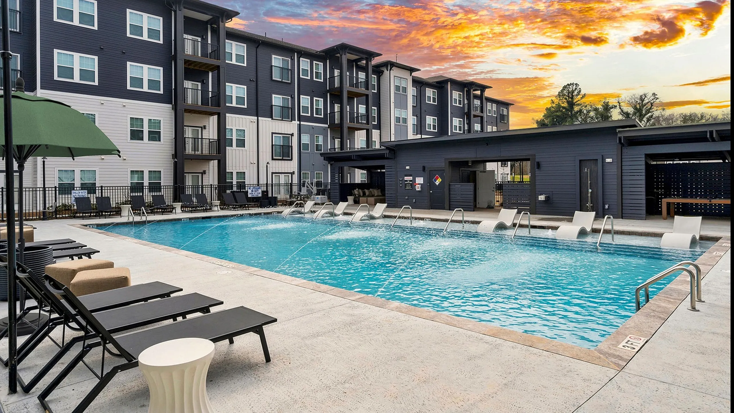 Stunning apartment complex with modern design, featuring a luxurious outdoor swimming pool and relaxing lounge chairs, set against a vibrant sunset sky.
