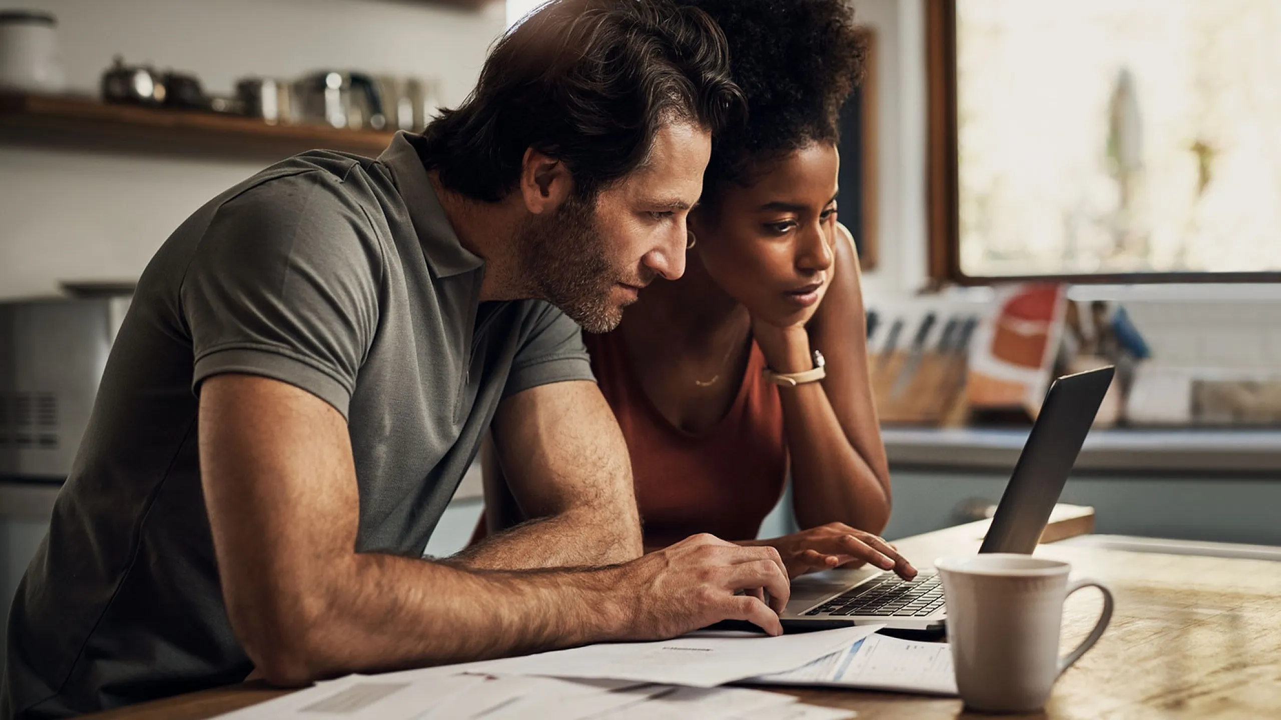 Focused couple in a cozy kitchen discussing plans on a laptop, surrounded by paperwork and enjoying a coffee.