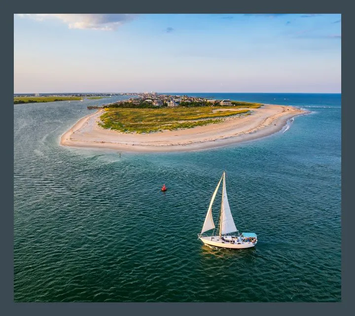 Stunning aerial view of a serene island surrounded by blue waters, with a graceful sailboat gliding nearby under a clear sky.