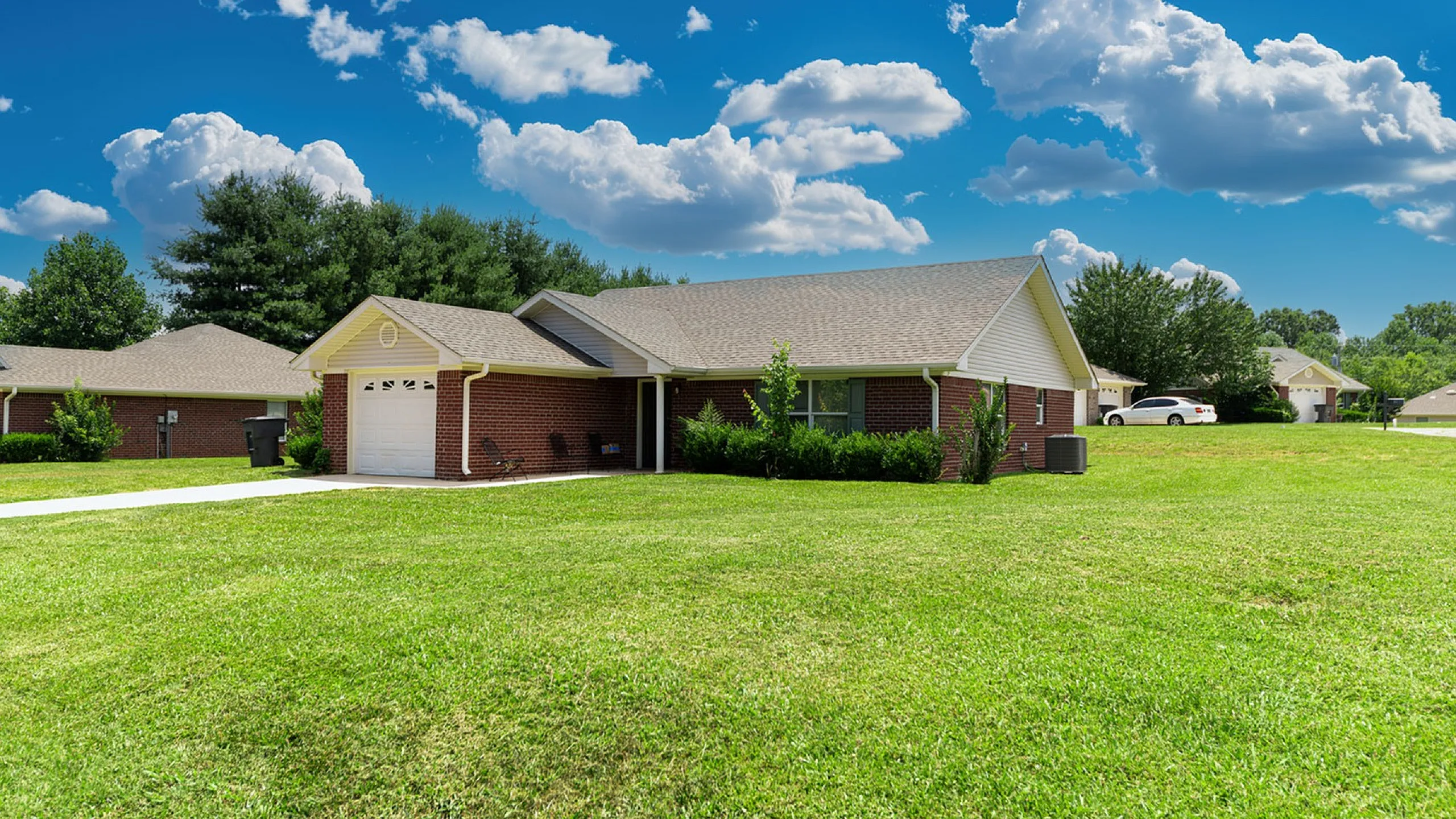 Charming brick home surrounded by lush green lawns under a bright blue sky with fluffy clouds.