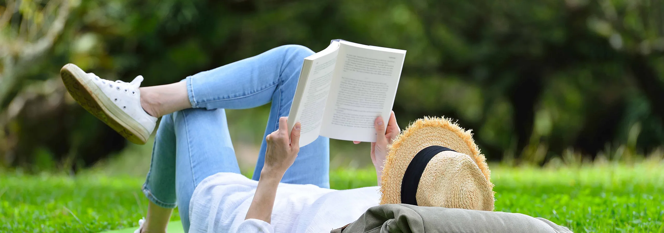 Peaceful outdoor scene with a person enjoying reading on a grassy lawn, wearing casual jeans and a straw hat.
