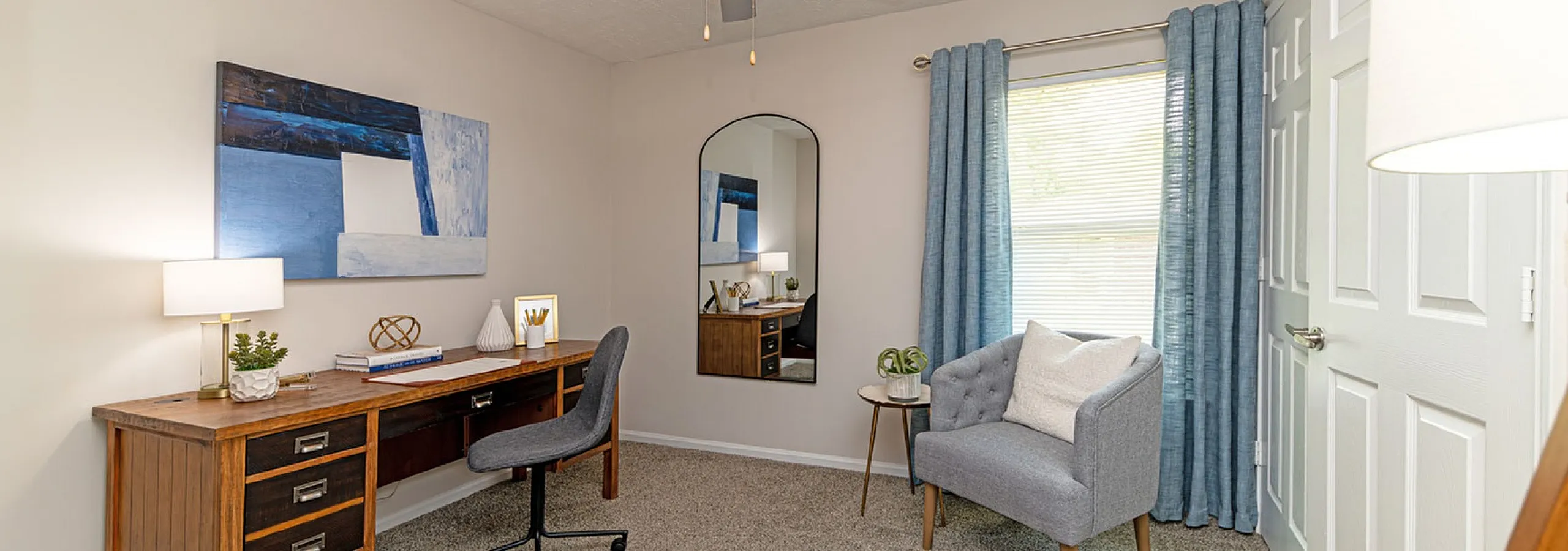 Charming home office with a sleek ceiling fan, wooden desk, and comfortable seating, bathed in natural light from large window with blue drapes.