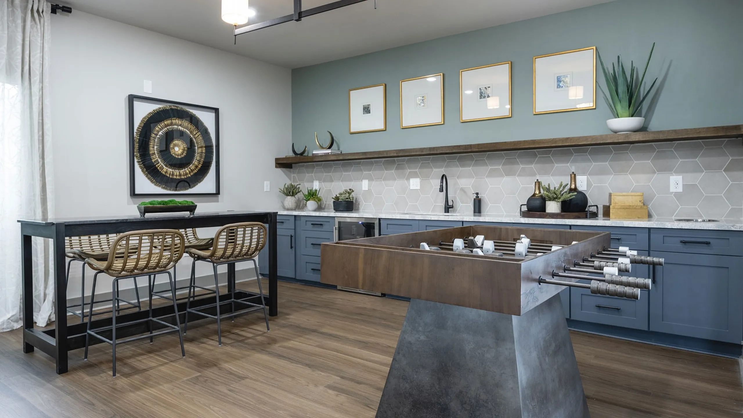 Chic apartment kitchen with modern blue cabinetry, elegant hexagonal backsplash, and stylish art pieces, complemented by a sleek breakfast bar and a contemporary foosball table.