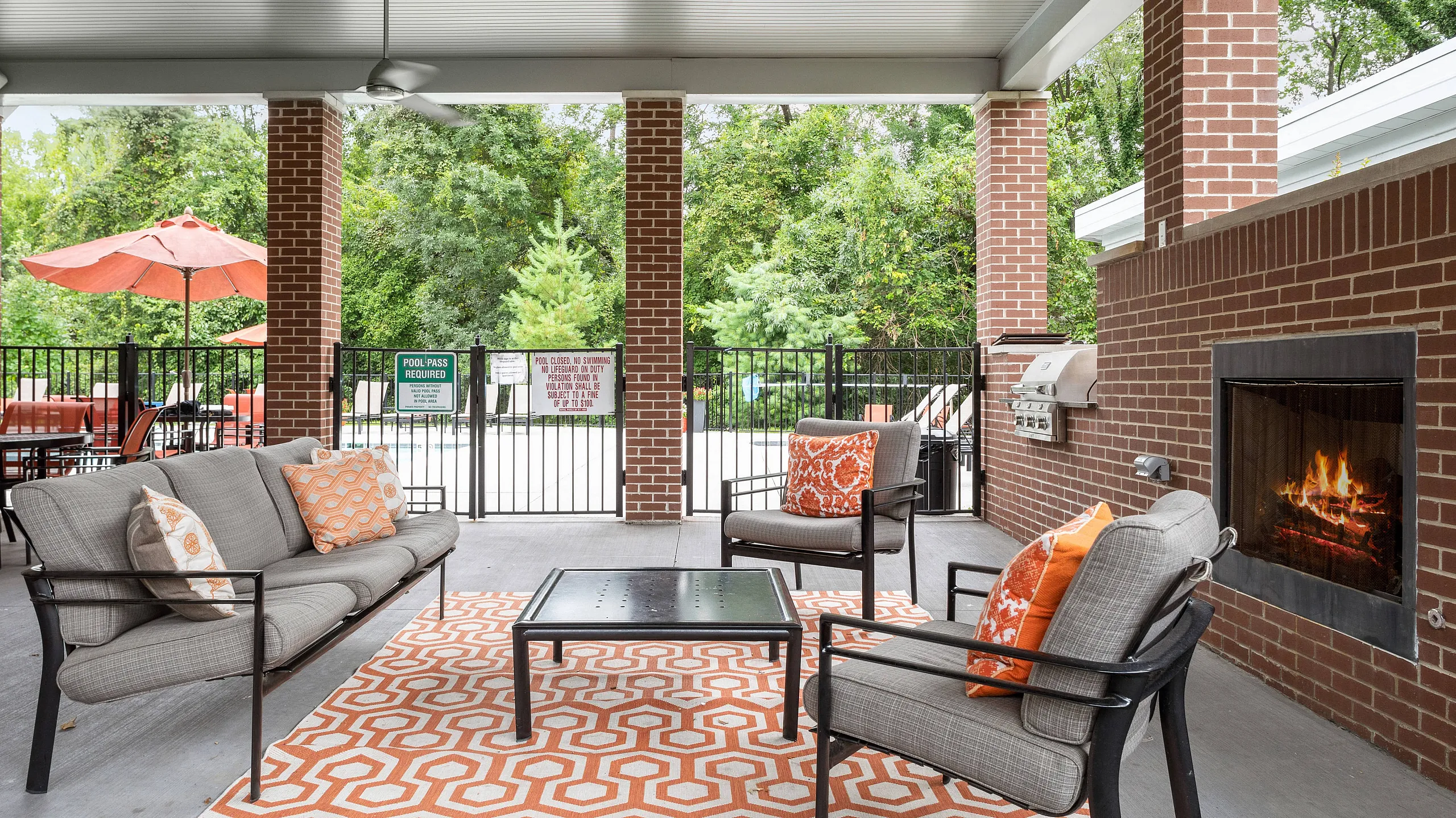 Charming outdoor patio with cozy seating, a brick fireplace, and vibrant orange accents, perfect for relaxing by the pool.