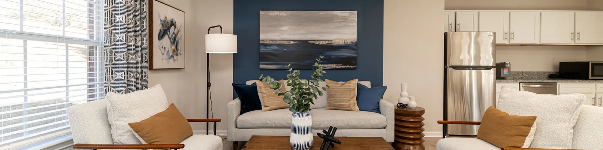 Elegant living room with modern furniture, cozy seating, stylish blue accents, and a sleek open kitchen in a bright, airy space.