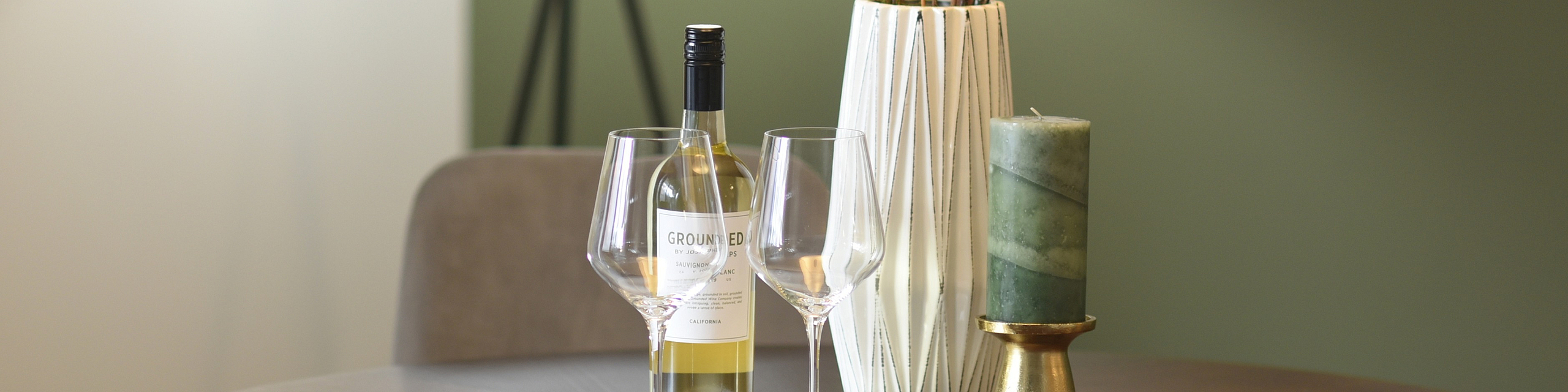 Cozy dining area with a stylish round table set for two, featuring a wine bottle, elegant glassware, and a chic centerpiece against a soft green wall.