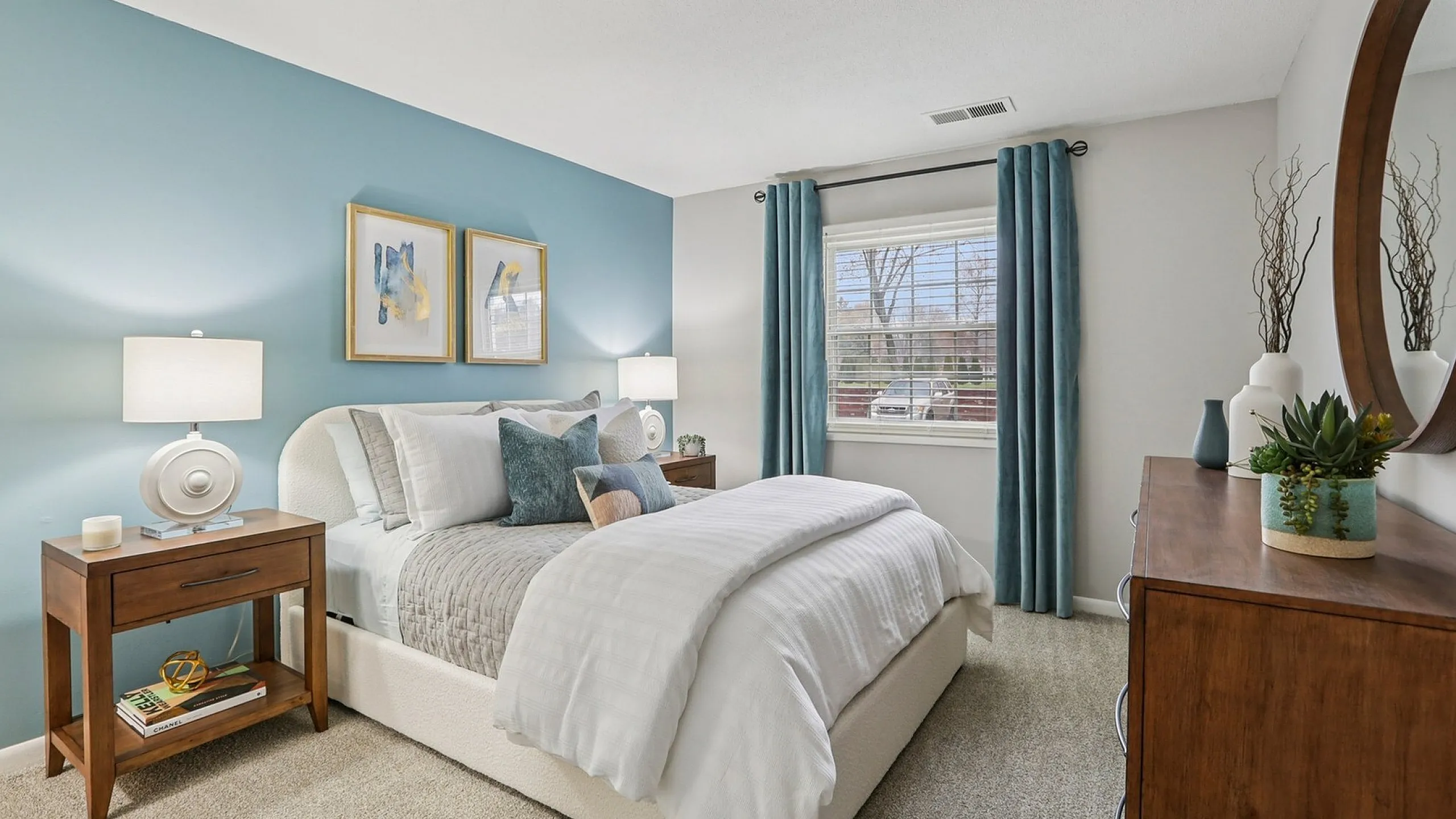 Elegant bedroom with a serene blue accent wall, featuring modern decor, plush bedding, and natural light from a large window.