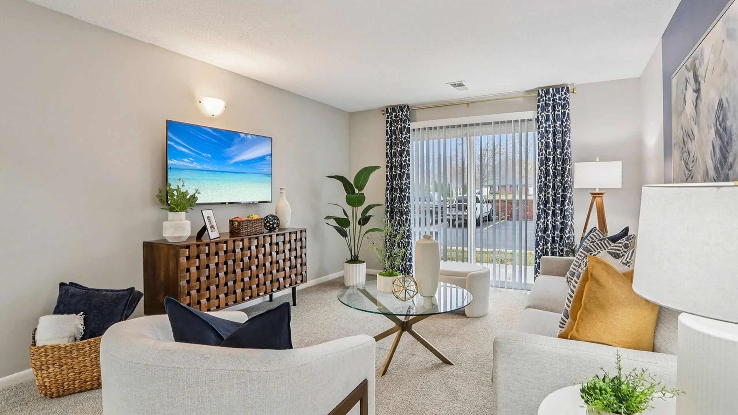 Bright and inviting living room with contemporary decor, featuring a sleek glass coffee table, plush chairs, and a flat-screen TV. Large windows with stylish drapes offer ample natural light and a serene outdoor view.