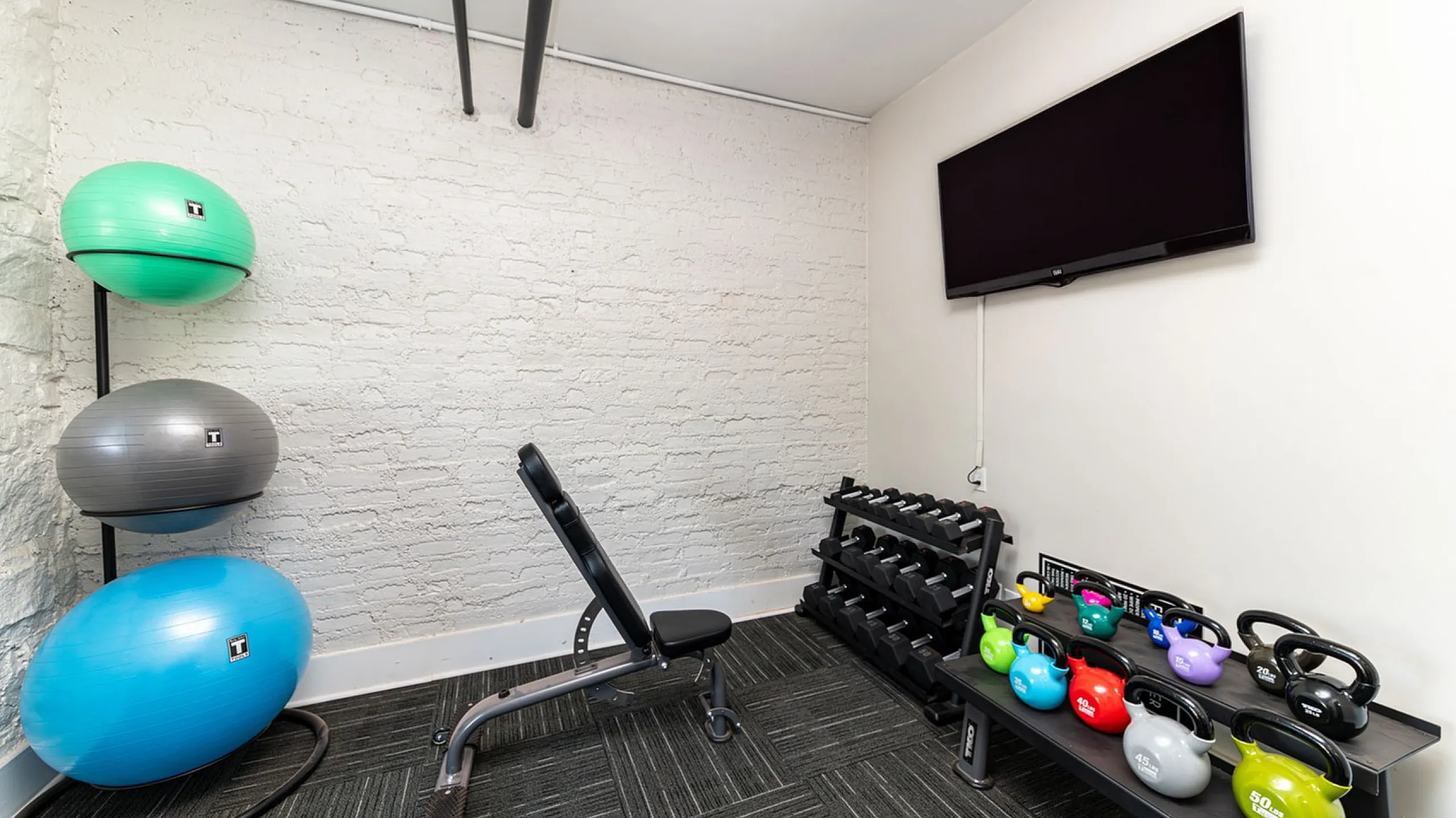 Chic home gym featuring a sleek bench, colorful kettlebells, exercise balls, and a modern TV for an enhanced workout experience.