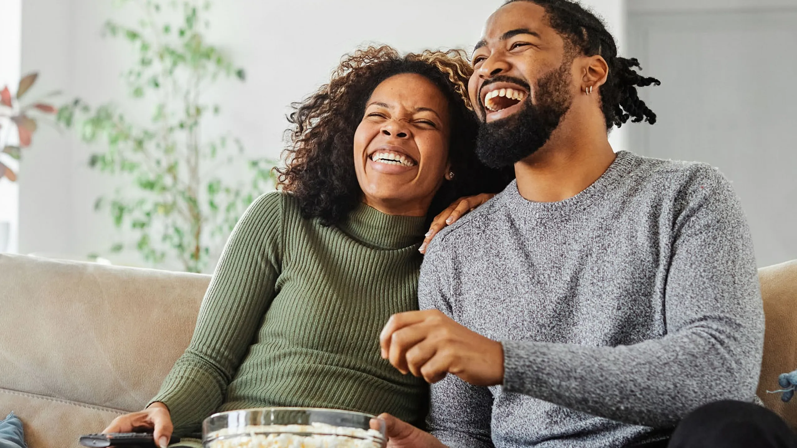 Joyful couple relaxing on a comfortable beige couch, enjoying a bowl of popcorn in a bright living room with lush green plants.