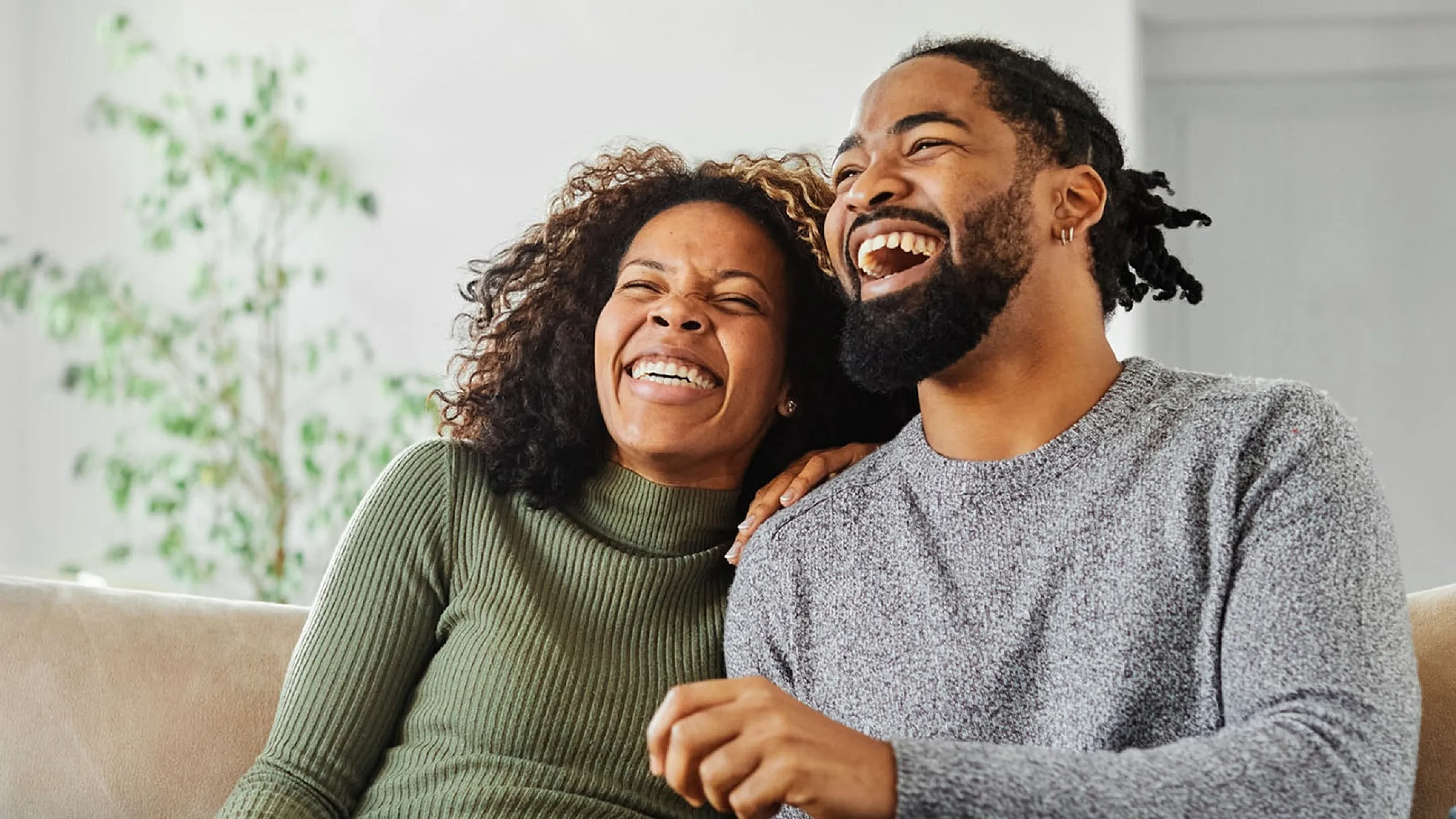 Cheerful couple enjoying a cozy moment on a modern beige sofa with a lush green plant in the background, creating an inviting and warm atmosphere.