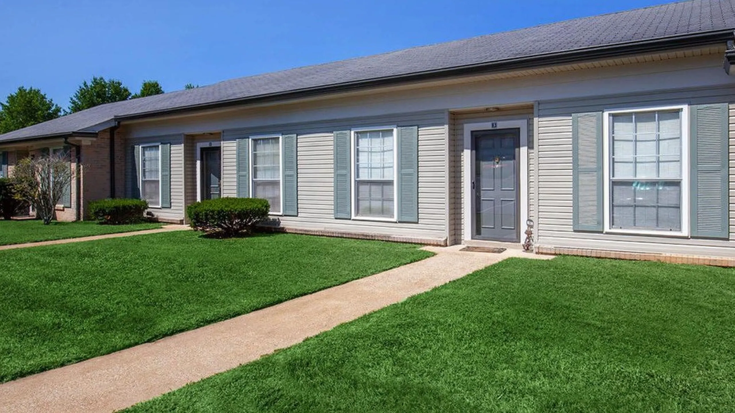 Charming single-story apartment building with elegant shutters, lush green lawns, and a welcoming sidewalk entry.