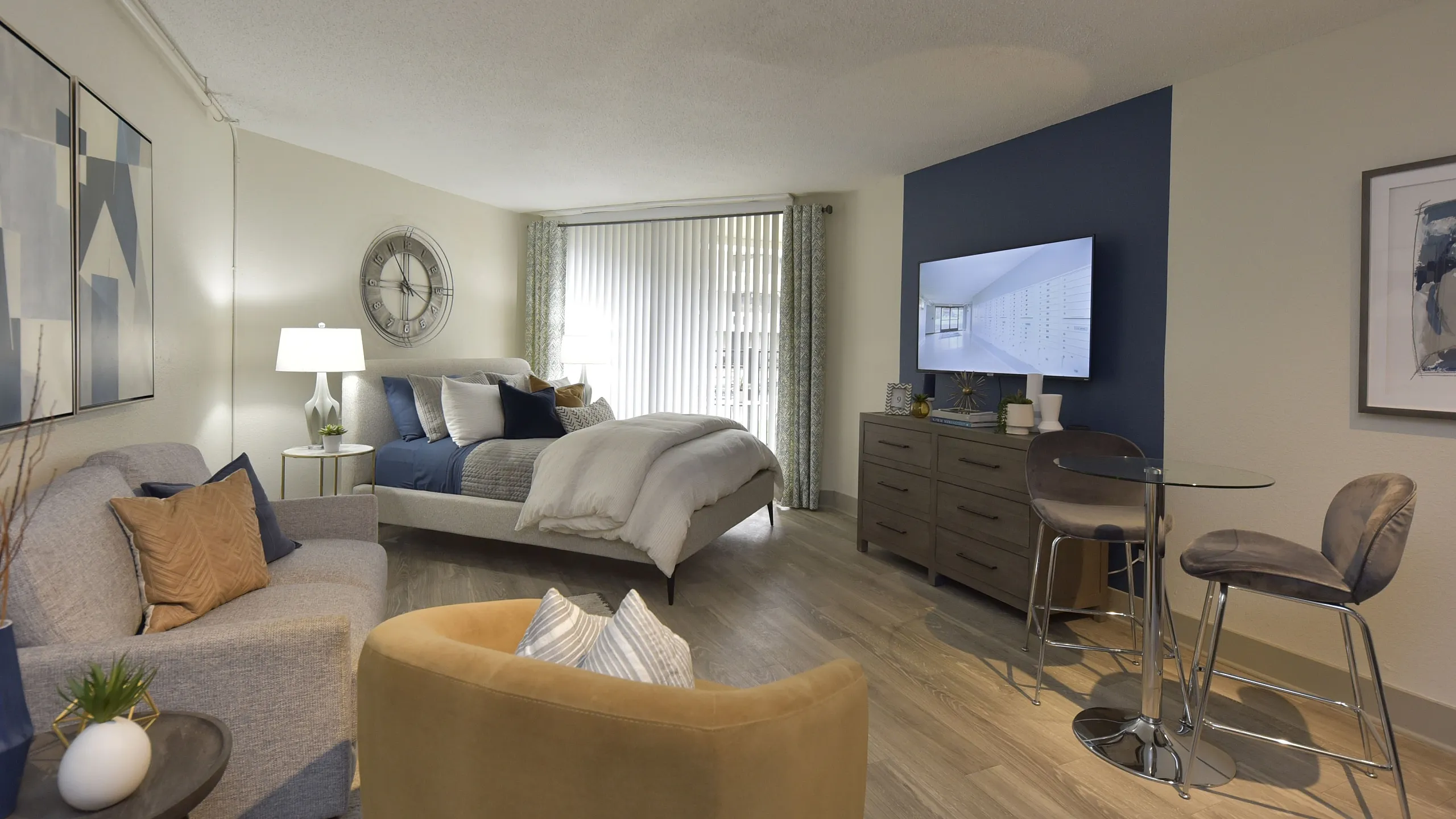 Modern studio apartment featuring a cozy sleeping area with a plush bed, stylish seating with accent chairs, a sleek dining space with a glass table, and elegant decor against a rich blue accent wall.