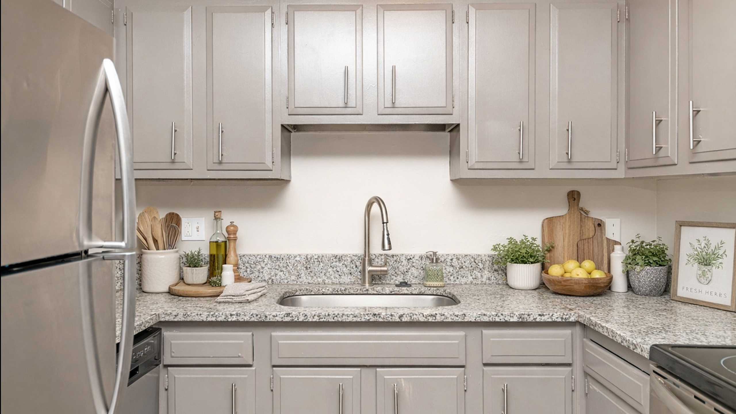 Modern kitchen with sleek gray cabinets, granite countertops, stainless steel appliances, and stylish decor elements like a wooden chopping board and fresh herbs.