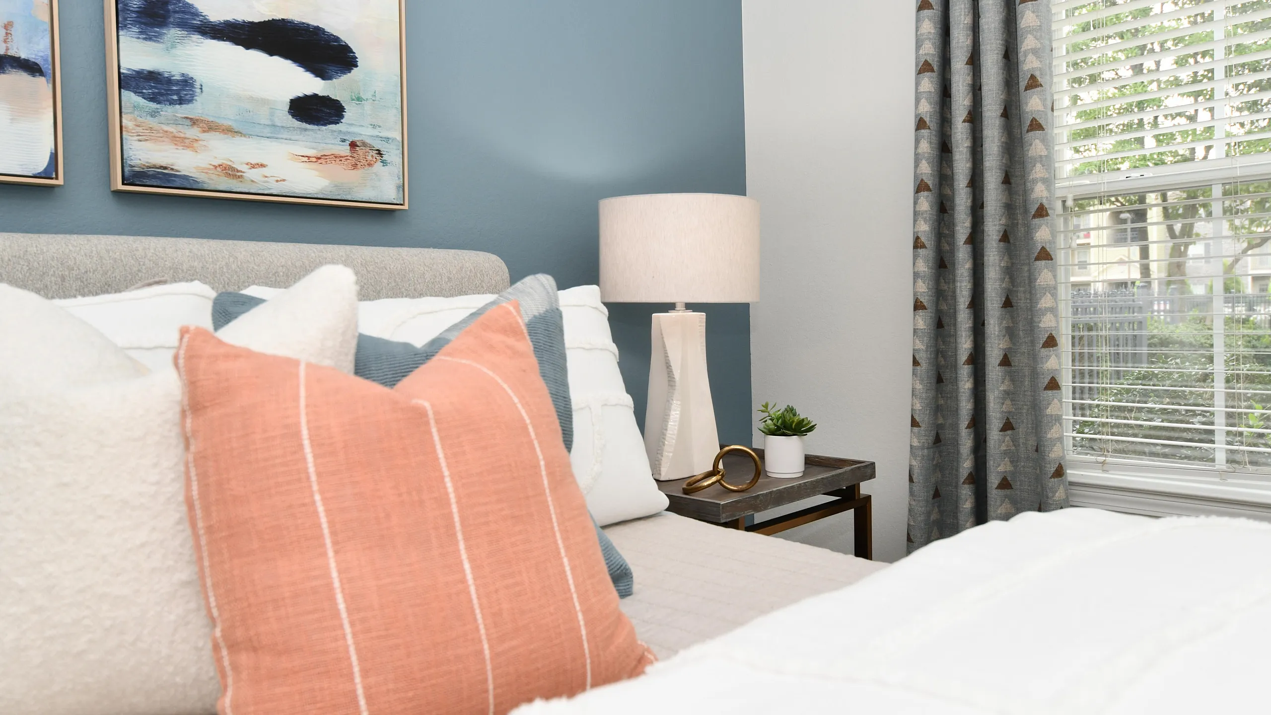 Chic bedroom with modern decor, featuring decorative pillows, abstract artwork, and elegant lighting against a calming blue accent wall.