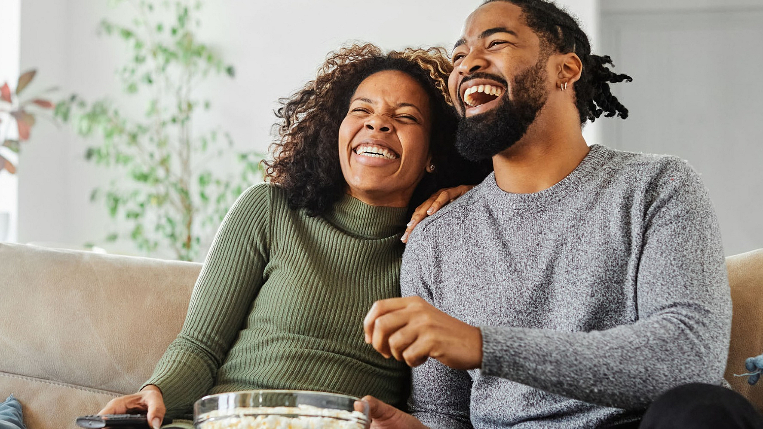 Joyful couple relaxing on a comfortable beige couch, enjoying a bowl of popcorn in a bright living room with lush green plants.