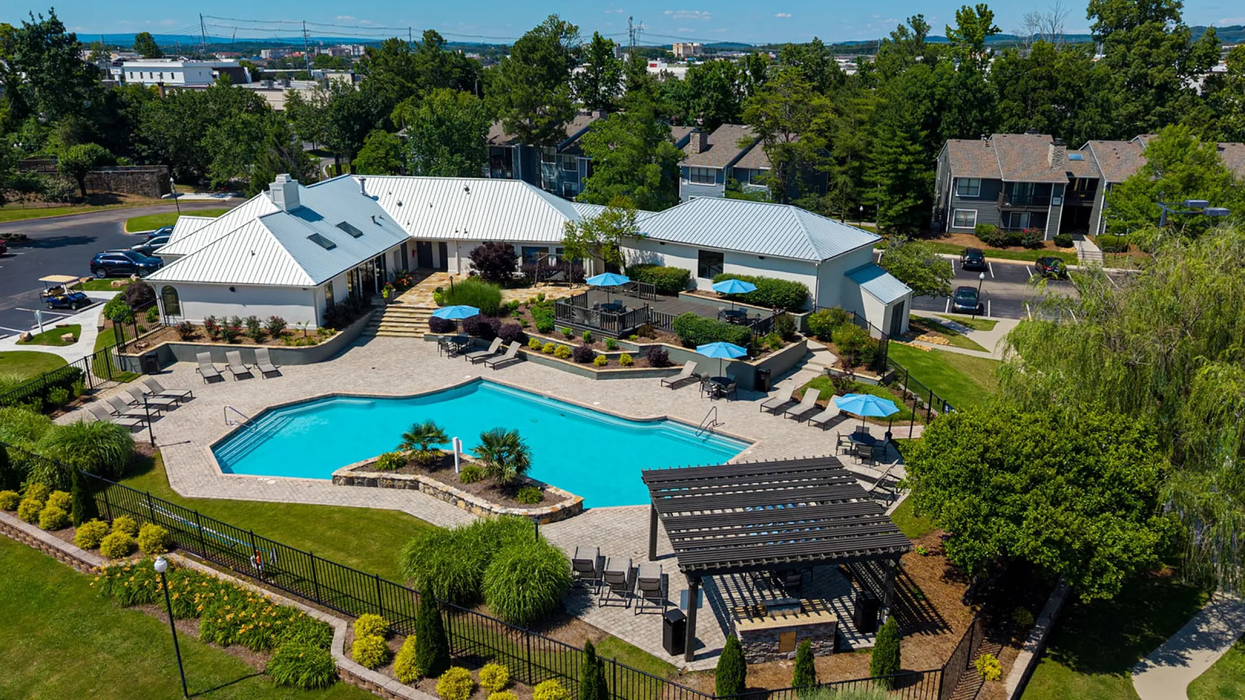Aerial view of a luxurious apartment complex featuring a modern clubhouse, expansive swimming pool, landscaped gardens, and cozy outdoor seating area.