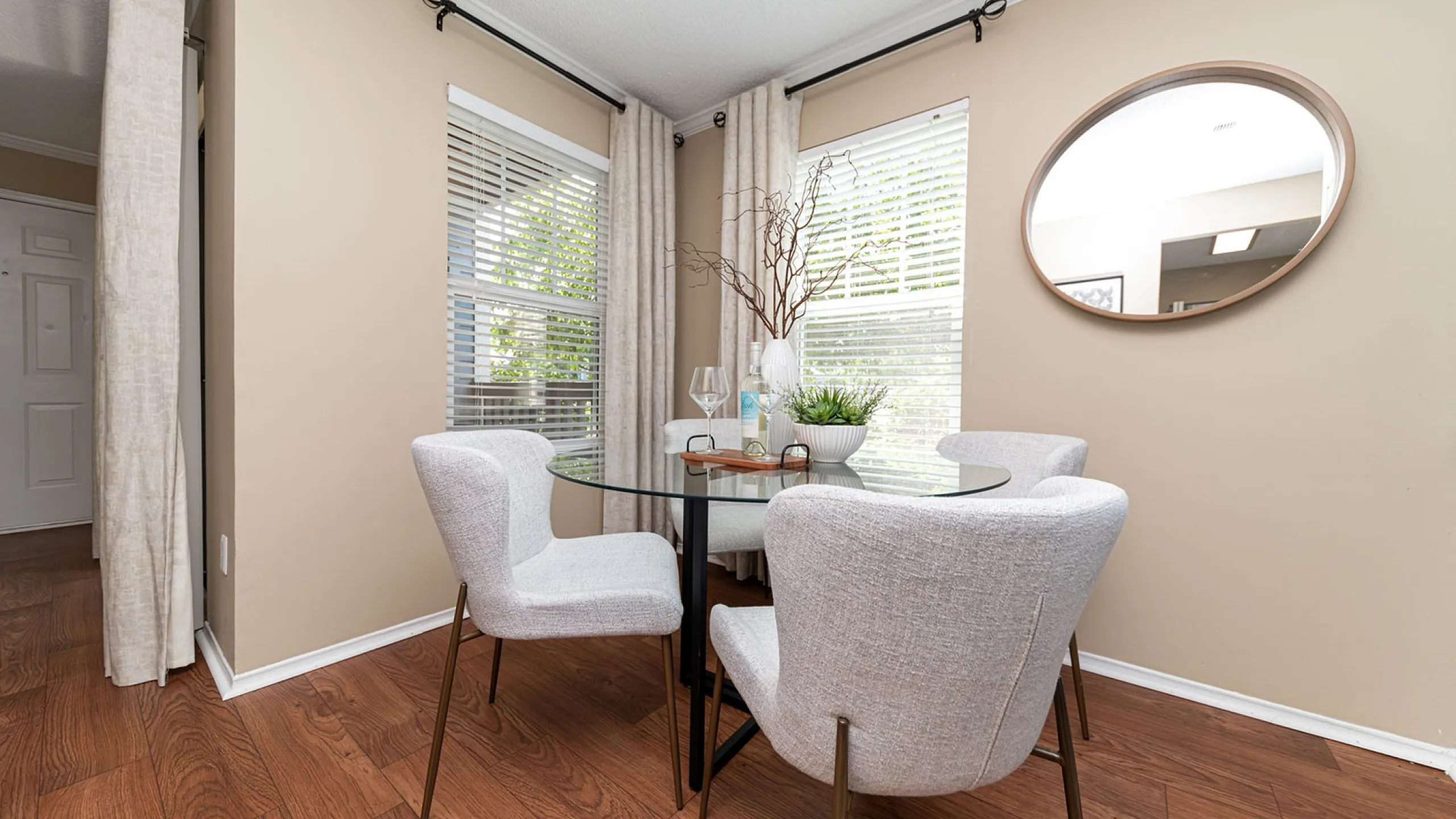 Elegant dining area with a round glass table, cushioned chairs, and tasteful decor beside large windows and a decorative mirror.