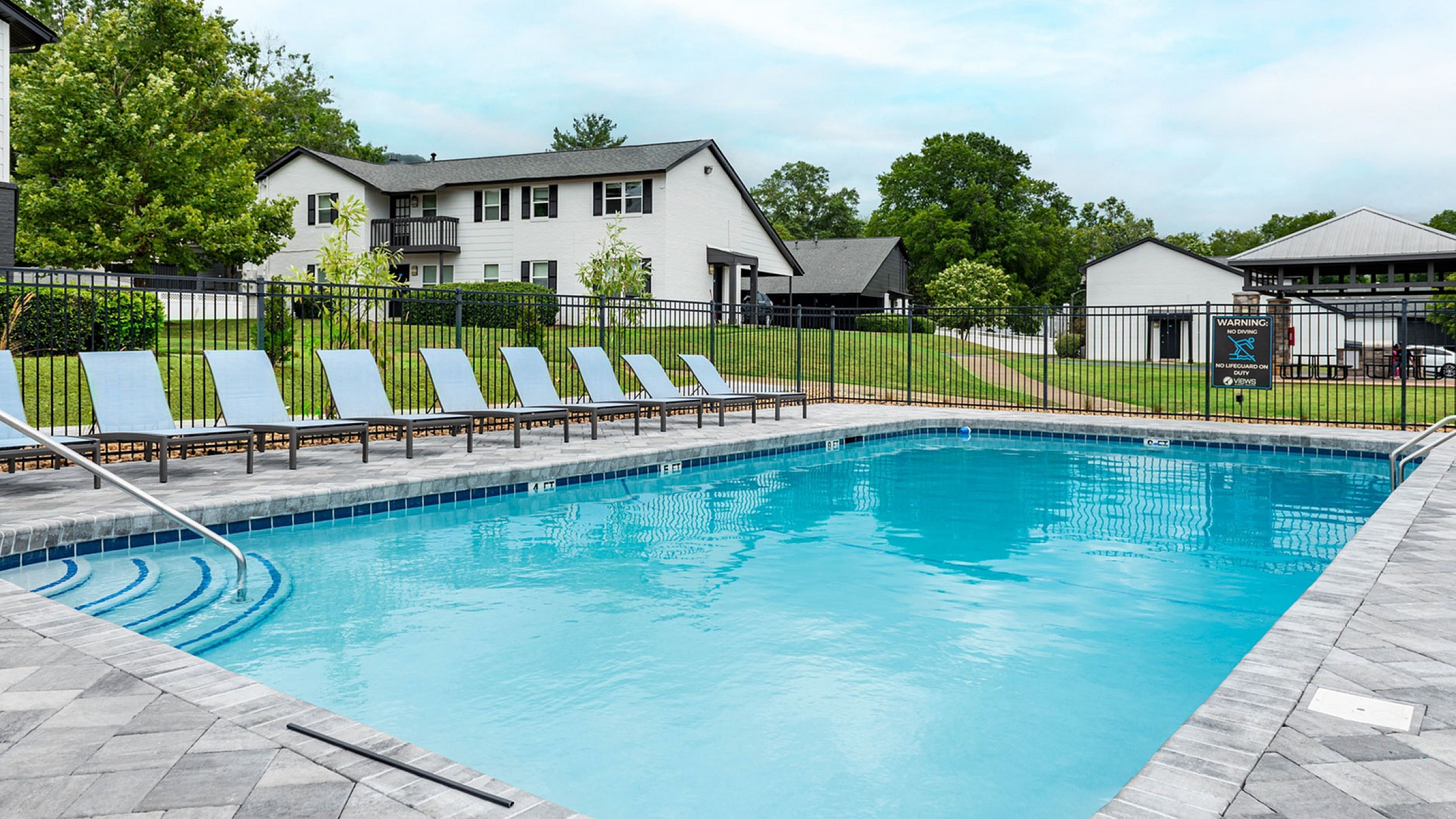 Inviting apartment complex with a pristine swimming pool surrounded by elegant lounge chairs, perfect for relaxation amidst a lush green landscape.