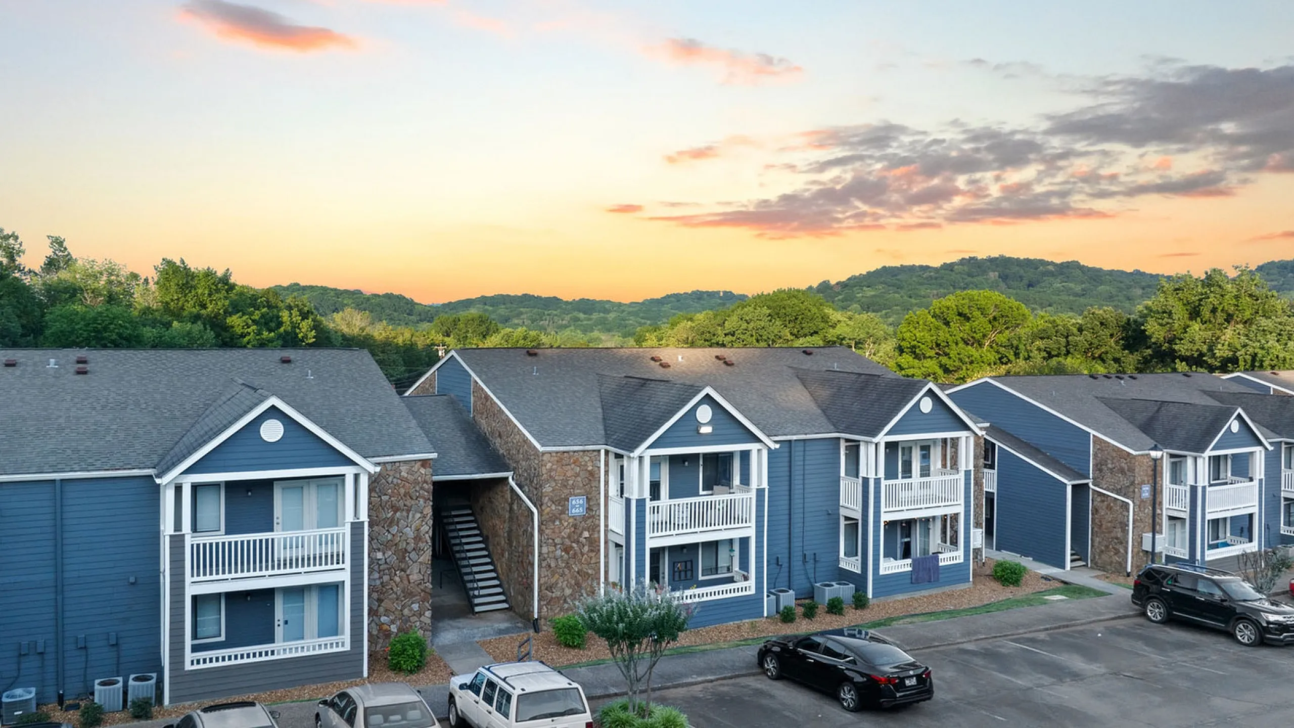 Charming apartment complex with modern blue siding, nestled against a backdrop of lush green hills and vibrant sunset skies.
