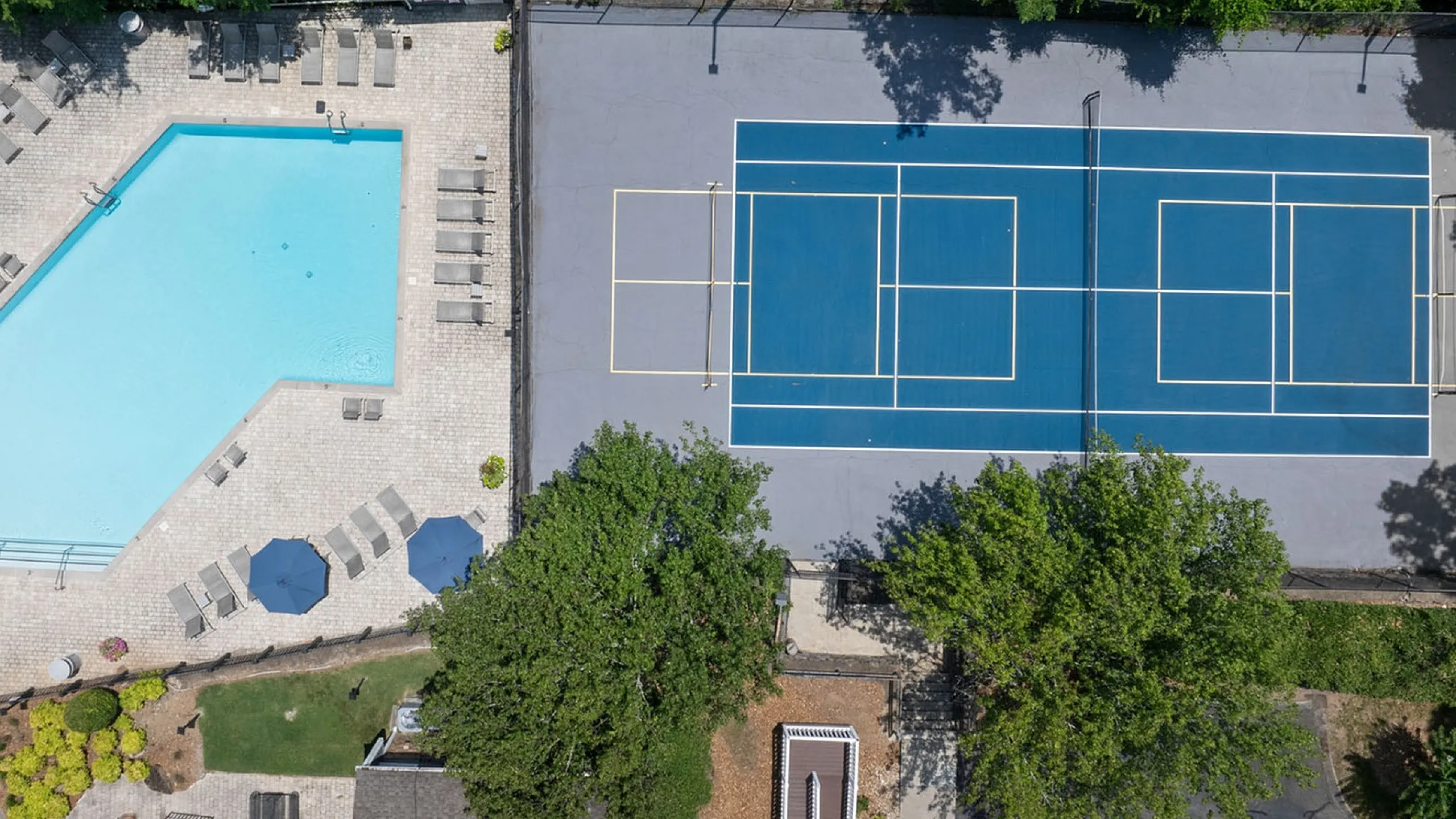 Aerial view of a vibrant recreational area featuring a sparkling swimming pool with lounge chairs and umbrellas, next to a well-maintained tennis court, surrounded by lush greenery.