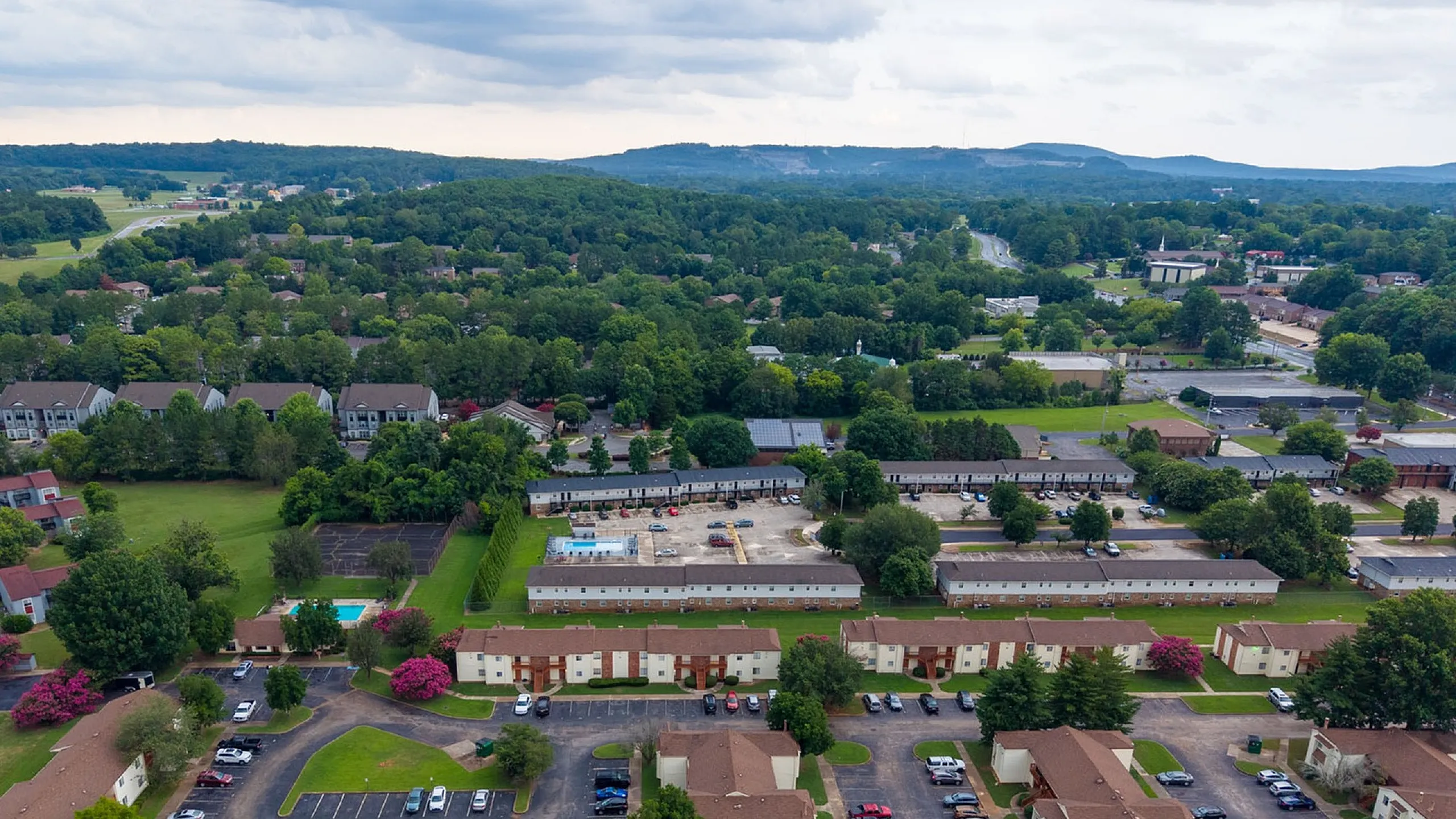 Charming aerial view of a lush apartment community surrounded by greenery, showcasing spacious parking and vibrant, landscaped grounds in a serene neighborhood.