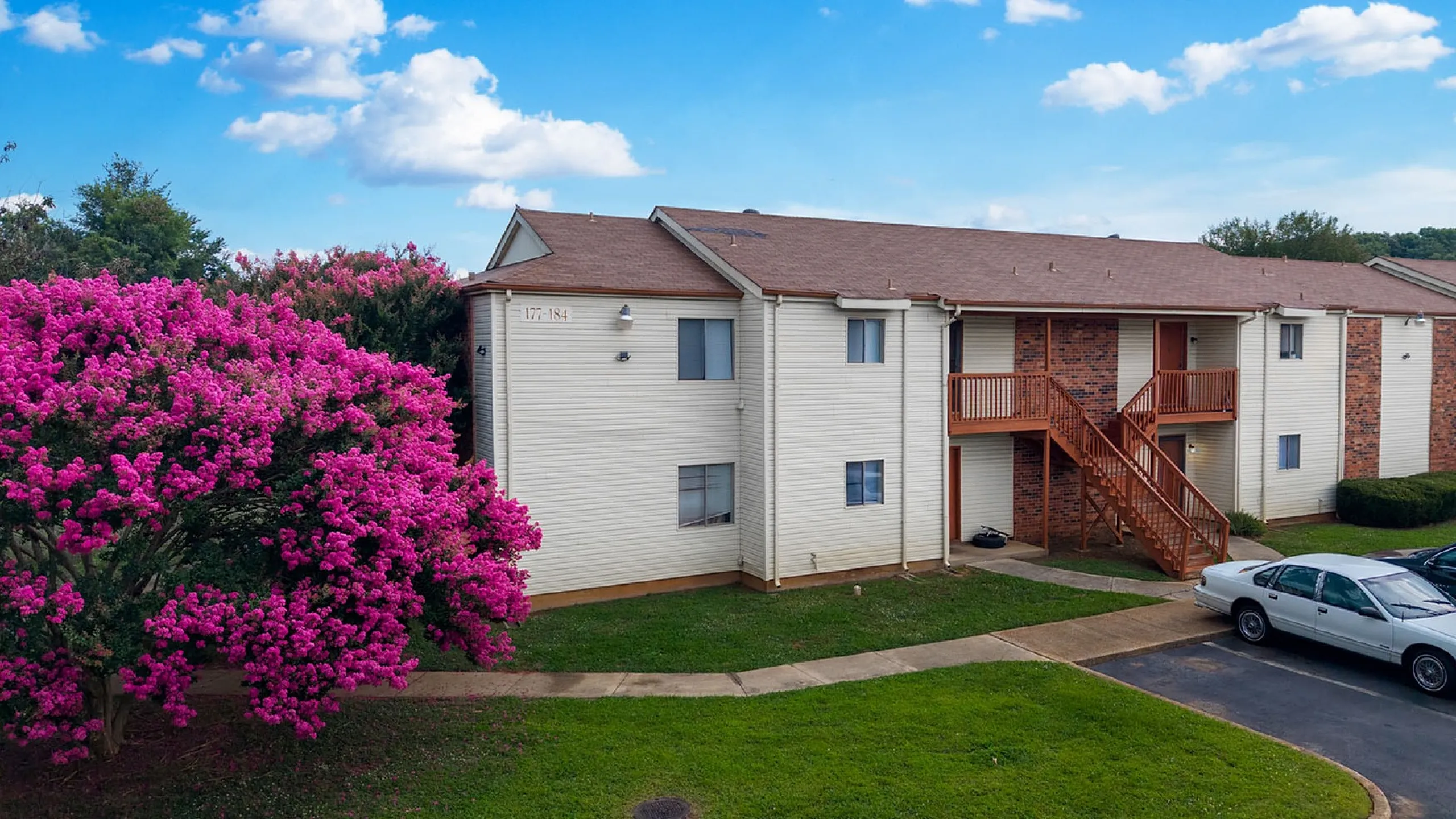 Charming apartment complex with a vibrant pink flowering tree against a bright blue sky, offering a welcoming entrance and convenient parking.