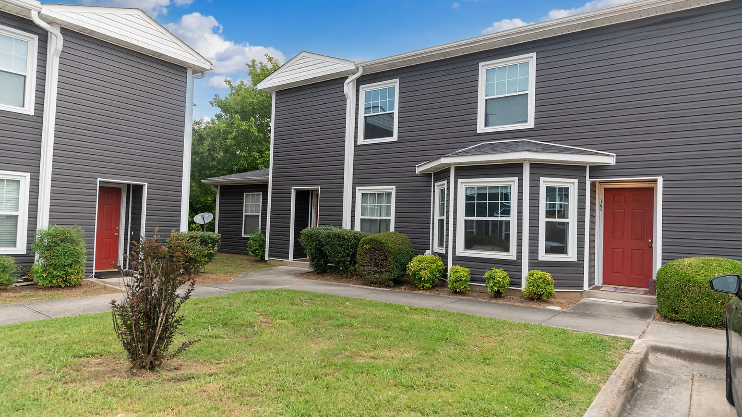 Charming townhouse with modern dark siding and vibrant red doors surrounded by lush greenery and clear blue skies, offering a welcoming neighborhood ambiance.