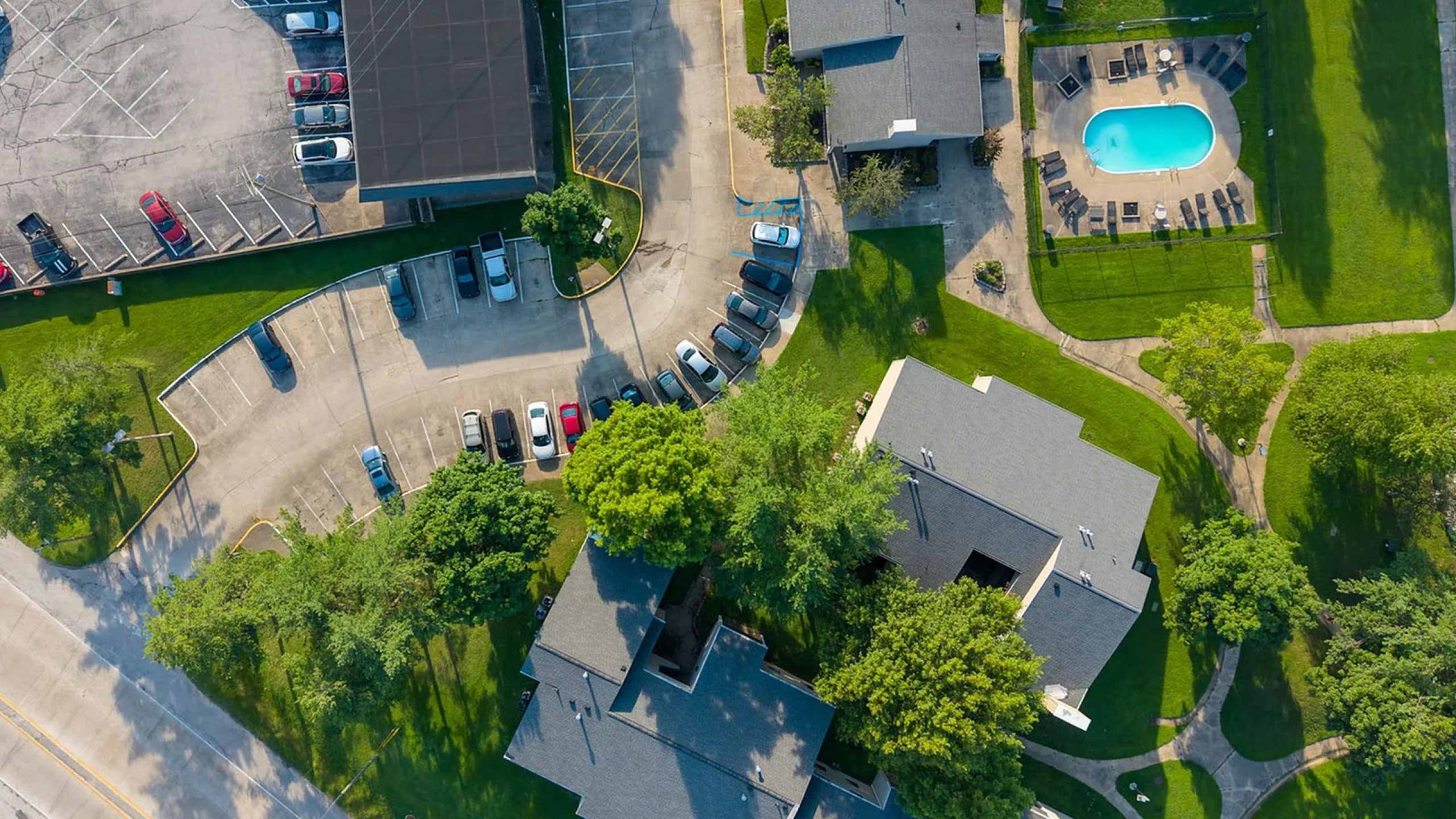 Aerial view of a vibrant apartment complex with lush green lawns, ample parking, and a sparkling blue swimming pool.