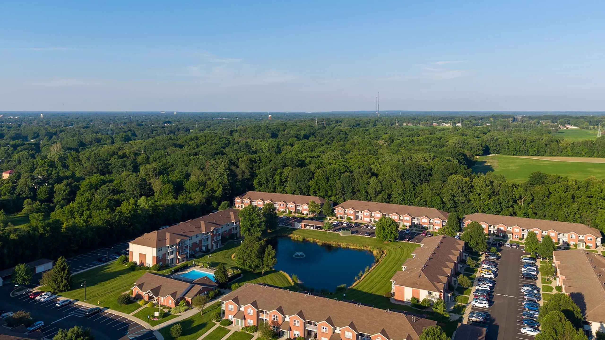 Scenic aerial view of apartment complex surrounded by lush greenery, showcasing community pool, pond, and ample parking.