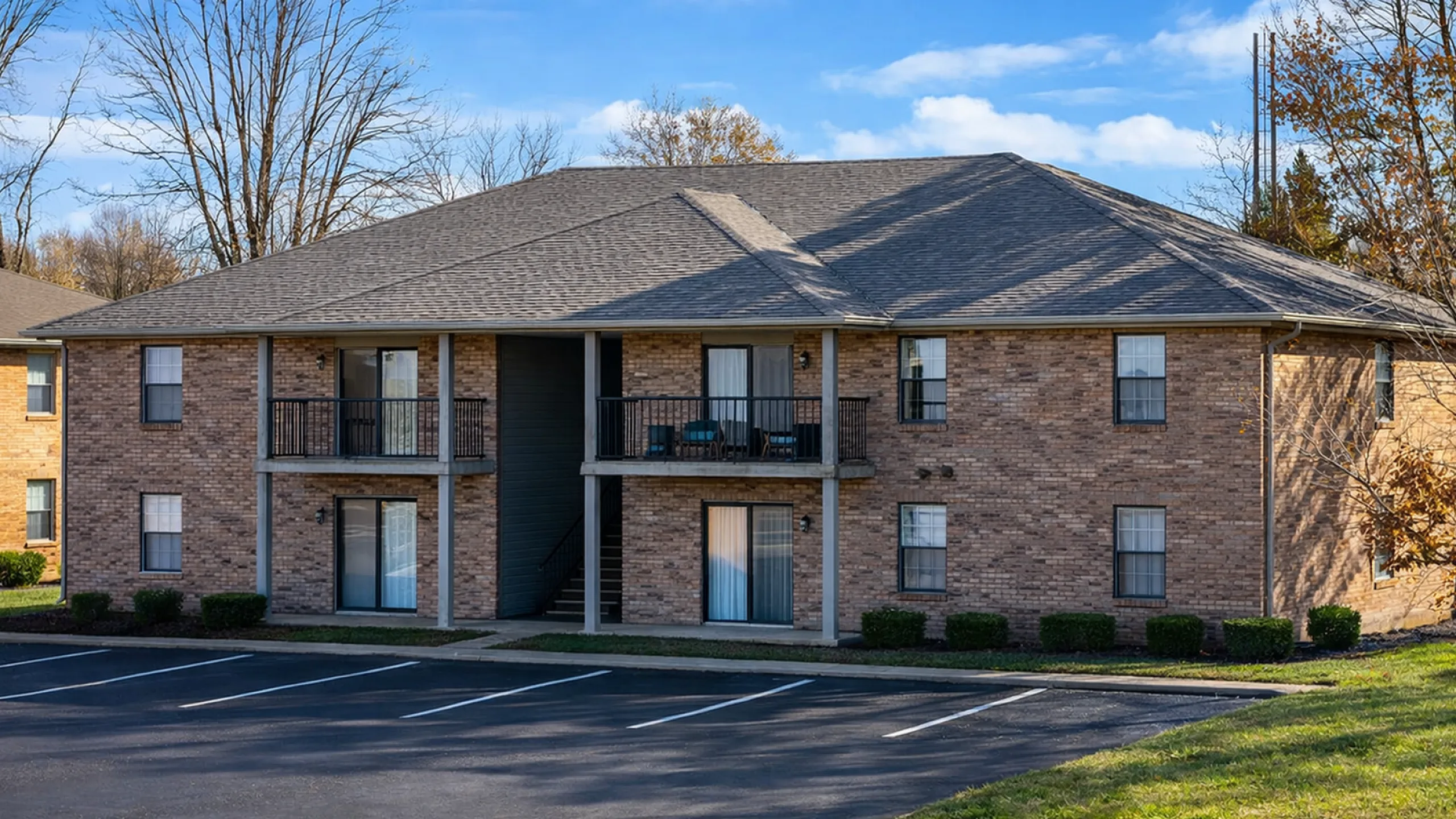 Charming brick apartment building with spacious balconies and a peaceful parking area, surrounded by vibrant greenery and a clear blue sky.