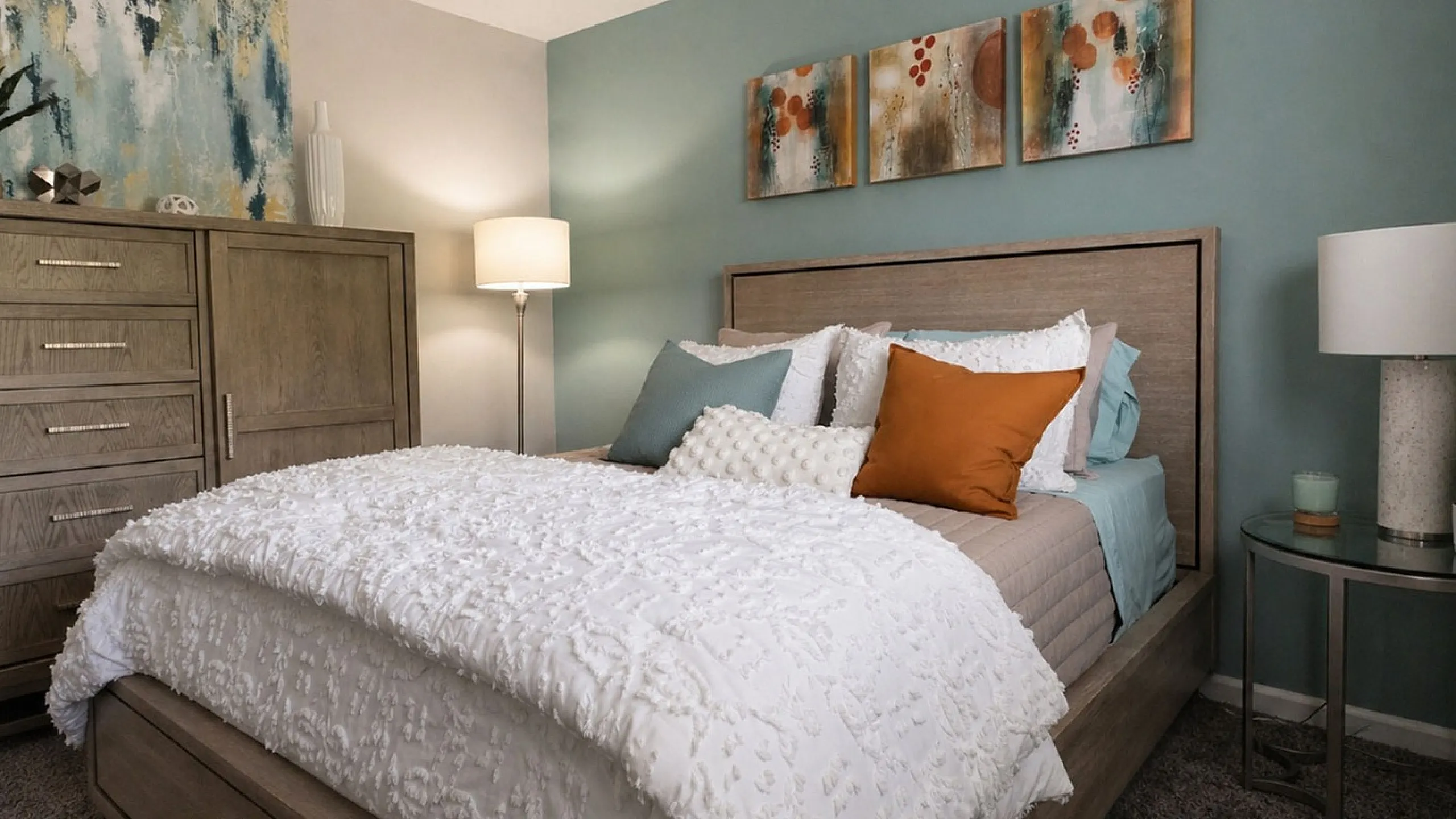 Chic bedroom with elegant decor, featuring a cozy bed with plush pillows, stylish dresser, and vibrant artwork on teal accent wall.